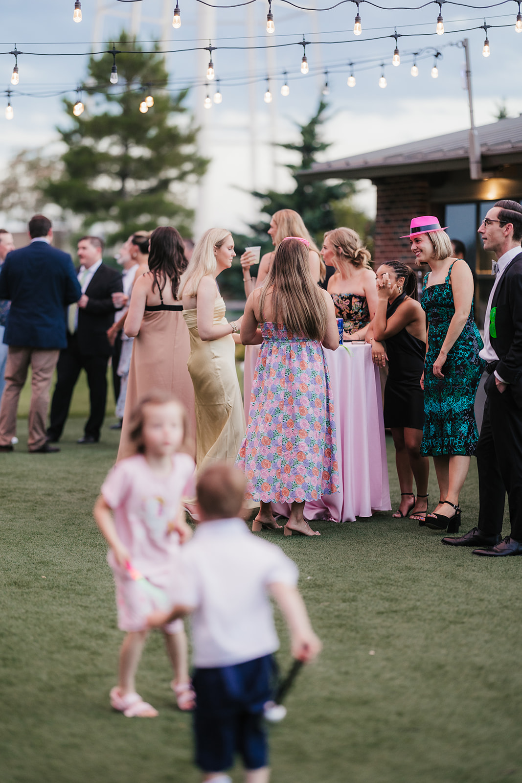 A group of adults in formal attire playfully interact with a young girl on a grassy lawn at an outdoor gathering.