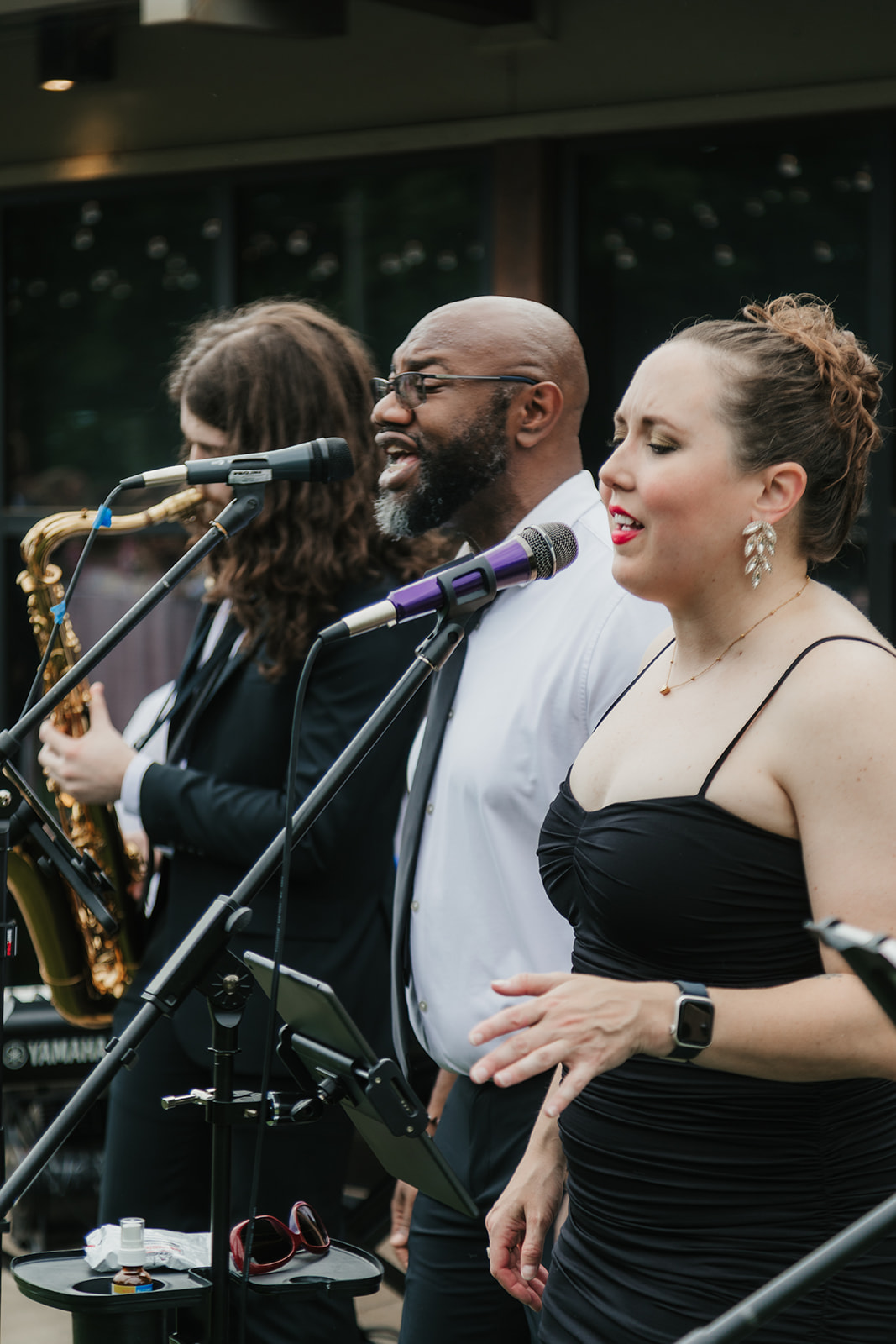 Three musicians perform outdoors: a saxophonist, a male vocalist, and a female vocalist in a black dress, each singing or playing into microphones.