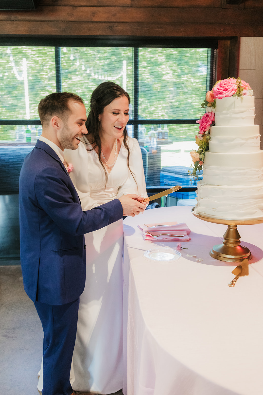 A bride and groom stand together, smiling as they cut a tiered white wedding cake decorated with pink flowers on a round table.
