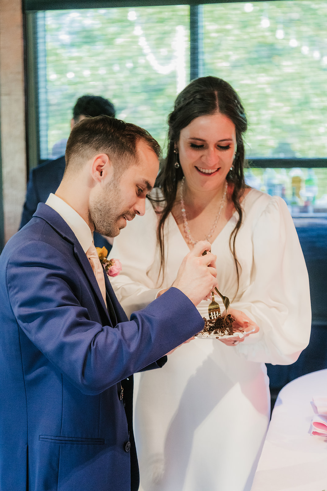 A bride and groom stand together, smiling as they cut a tiered white wedding cake decorated with pink flowers on a round table.