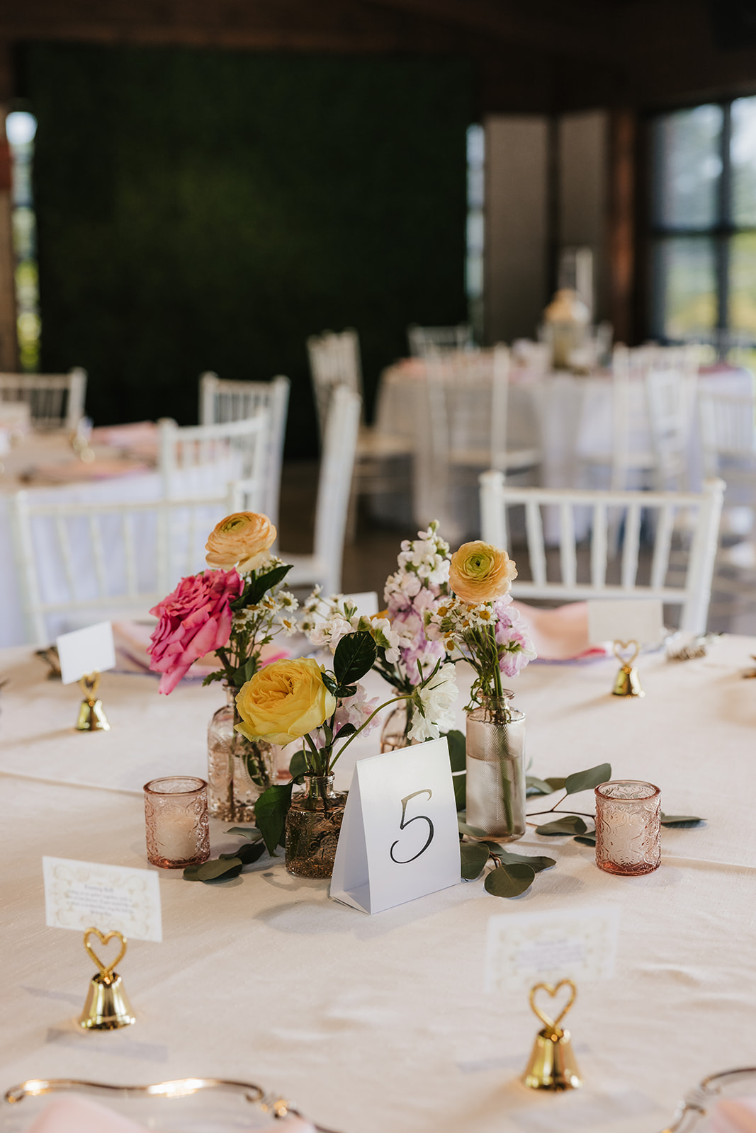 Round banquet table with white cloth, floral centerpiece in glass jars, table number 5, gold bell place card holders, and white chairs in a softly lit event setting.