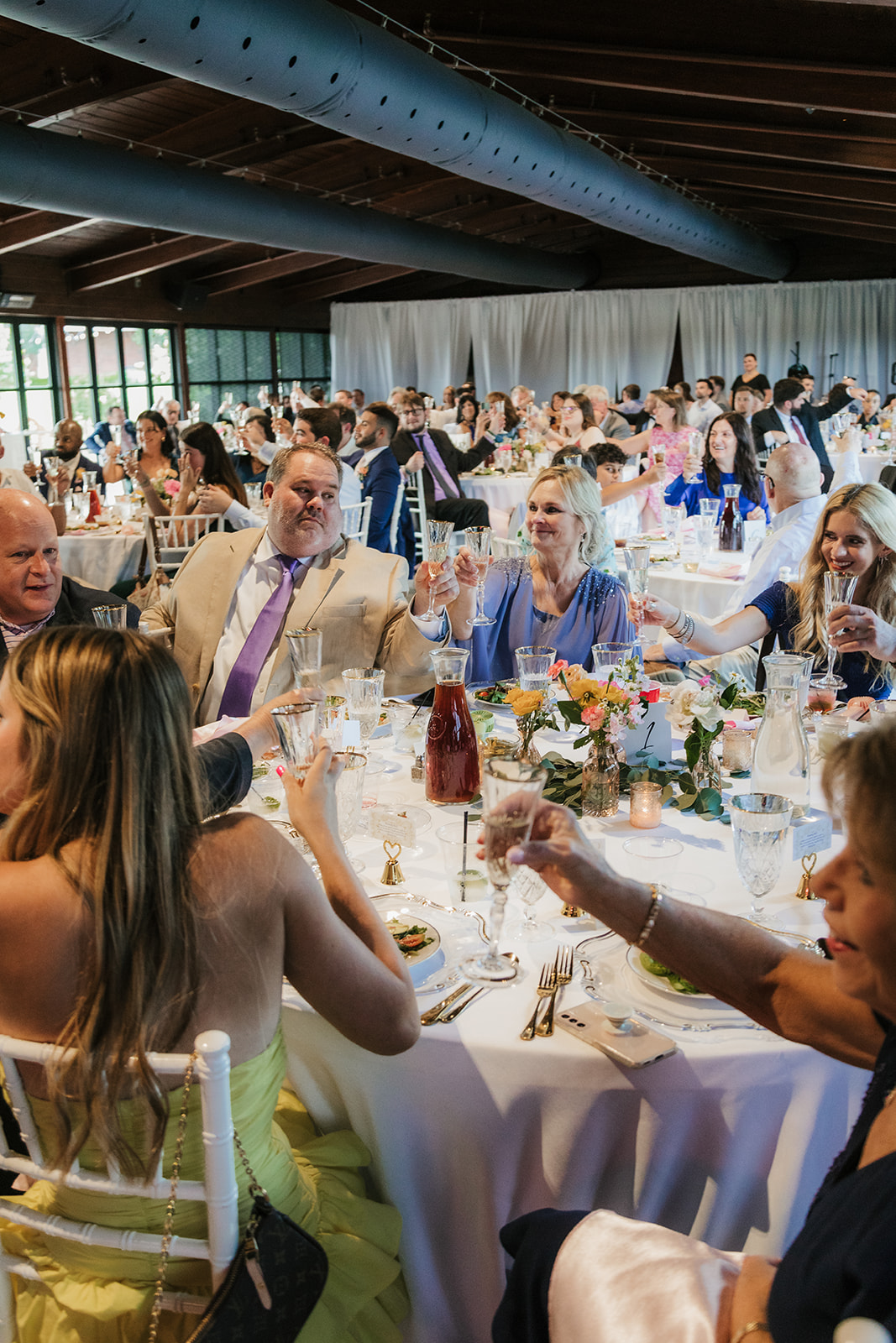 Guests seated at round tables raise their glasses in a toast during a formal event or celebration in a large, decorated indoor venue.