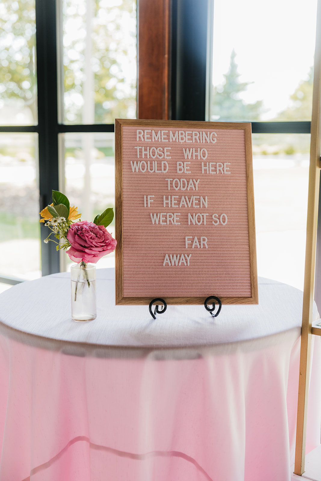 A sign on a table reads, "Remembering those who would be here today if heaven were not so far away," next to a vase with pink and yellow flowers.