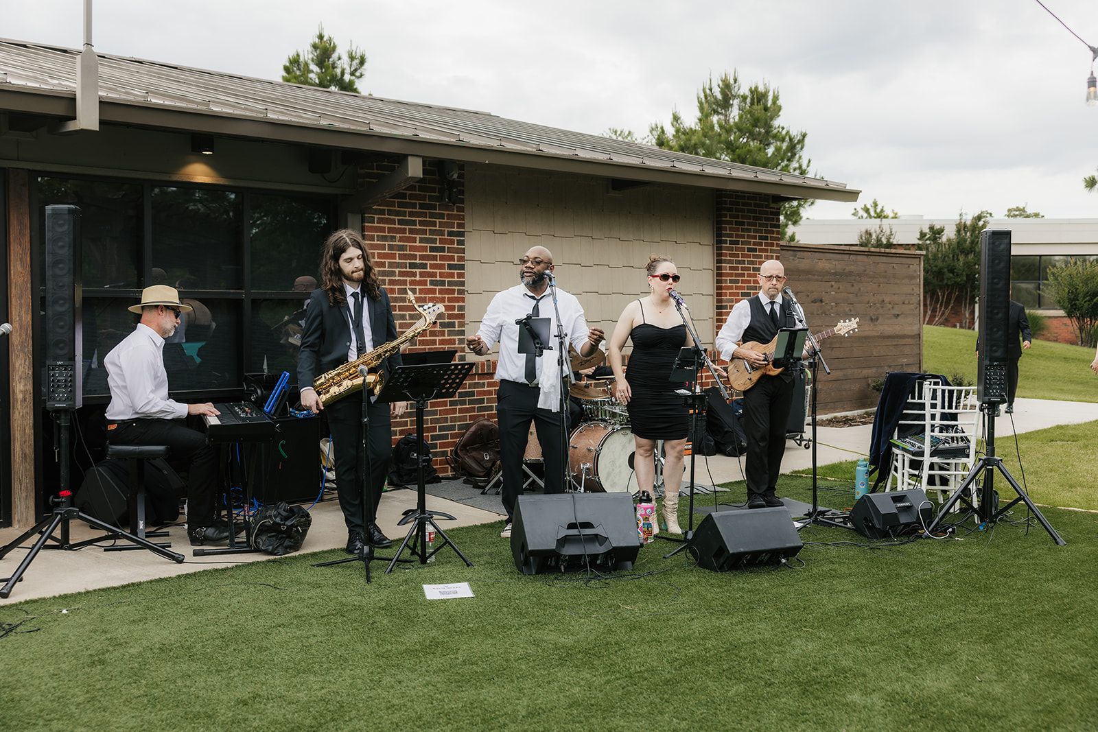 A five-member band performs outdoors on grass, with various instruments including keyboard, saxophone, guitar, and microphones, in front of a brick building.