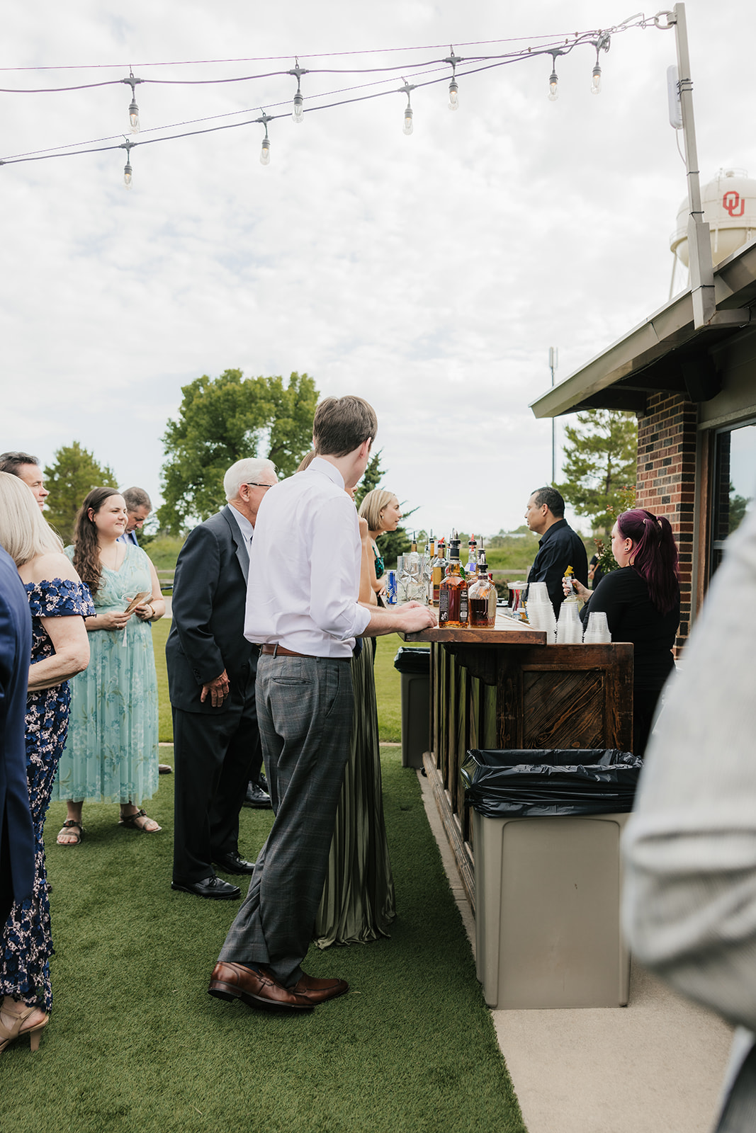 People stand in line at an outdoor bar under string lights, with trees and a cloudy sky in the background.
