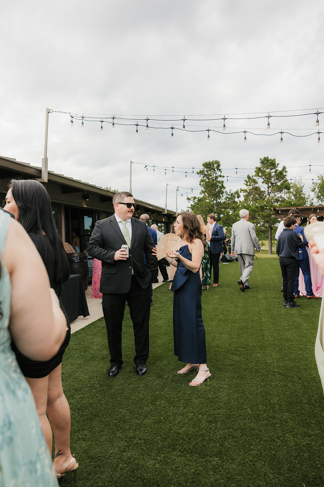 People stand in line at an outdoor bar under string lights, with trees and a cloudy sky in the background.
