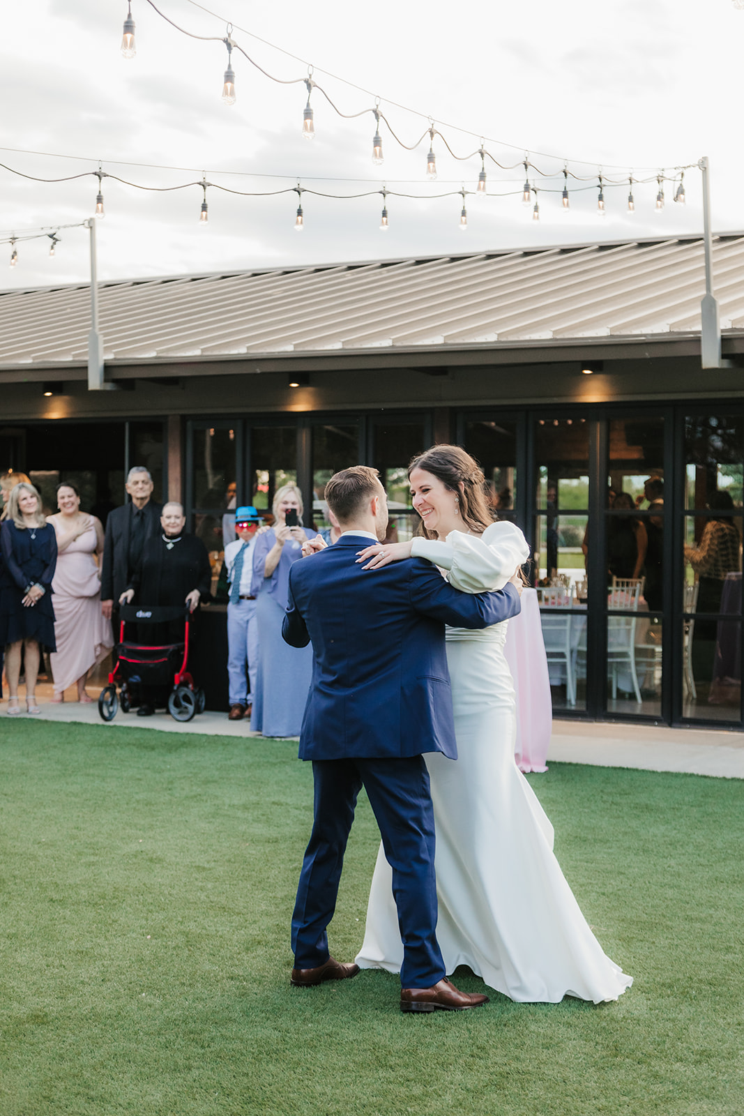 A bride and groom dance together outdoors on a lawn, with guests watching in the background near a building decorated with string lights.