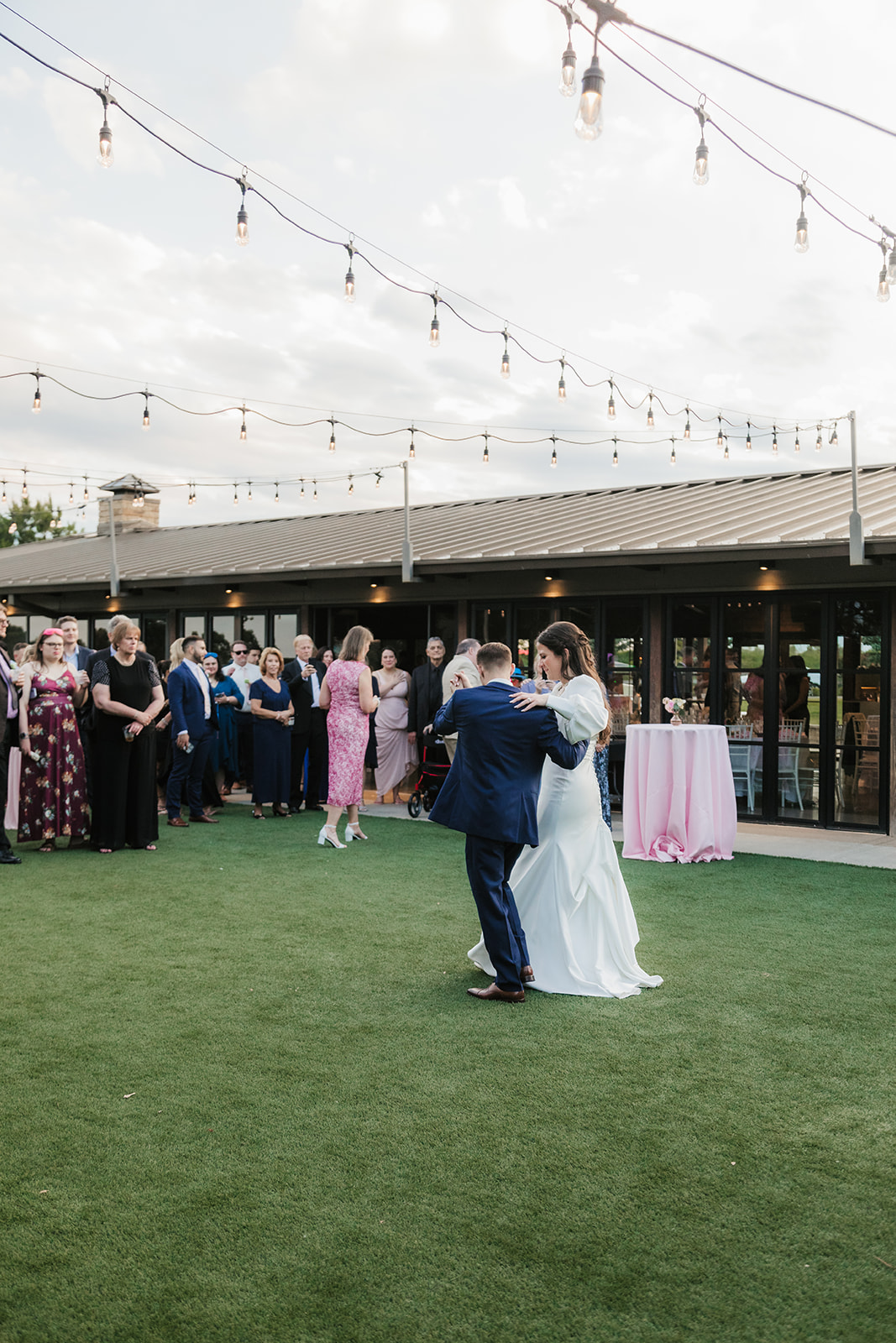 A bride and groom dance together outdoors on a lawn, with guests watching in the background near a building decorated with string lights.