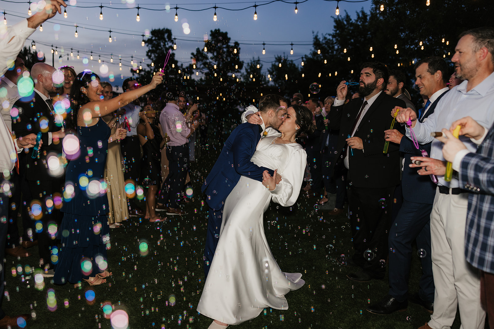 A bride in a white dress walks outdoors with her arms raised, surrounded by guests blowing bubbles at a nighttime wedding celebration.