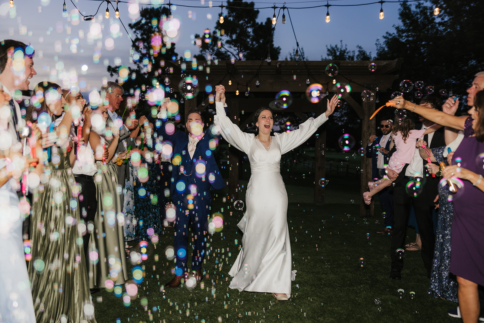 A bride in a white dress walks outdoors with her arms raised, surrounded by guests blowing bubbles at a nighttime wedding celebration.