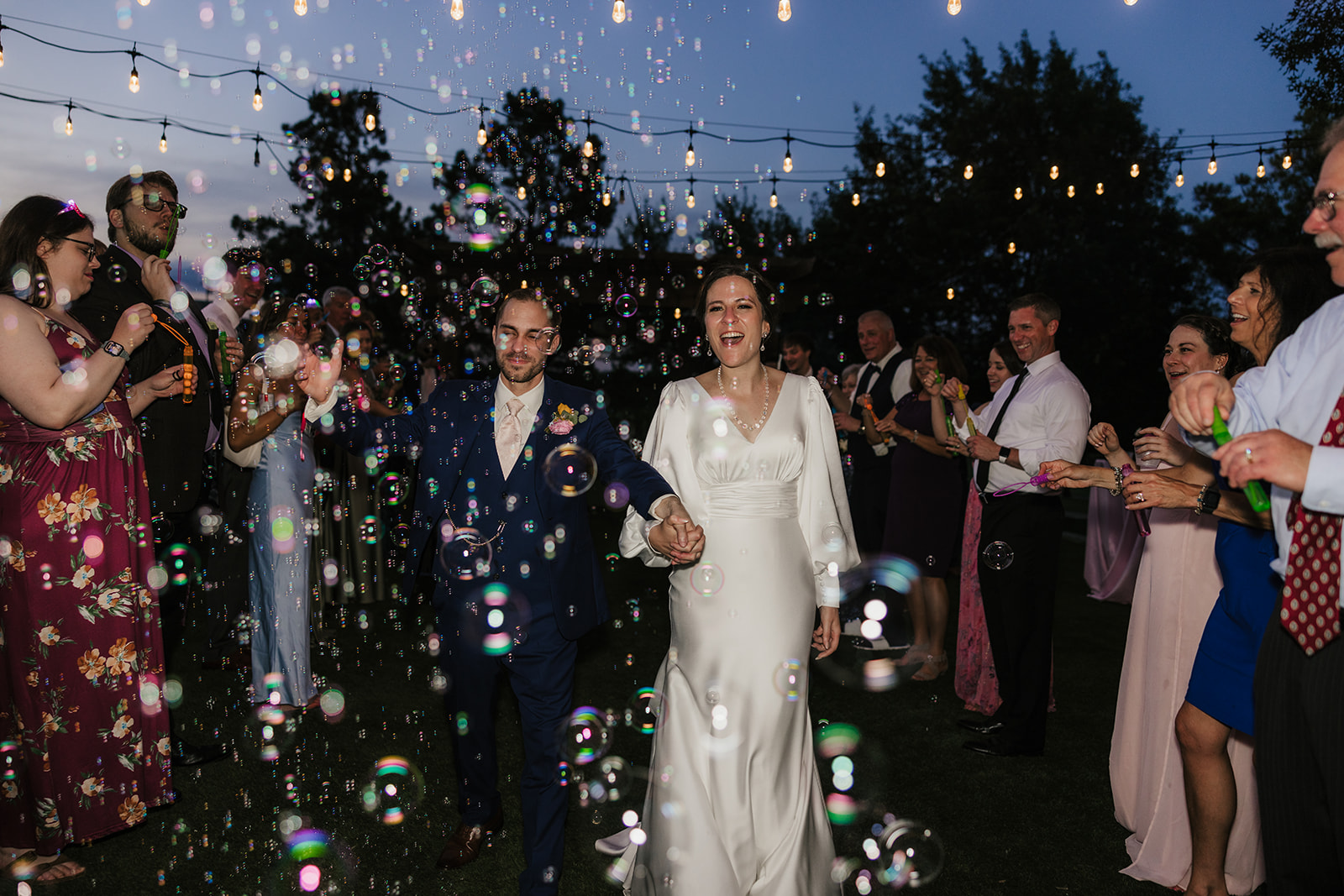A bride in a white dress walks outdoors with her arms raised, surrounded by guests blowing bubbles at a nighttime wedding celebration.