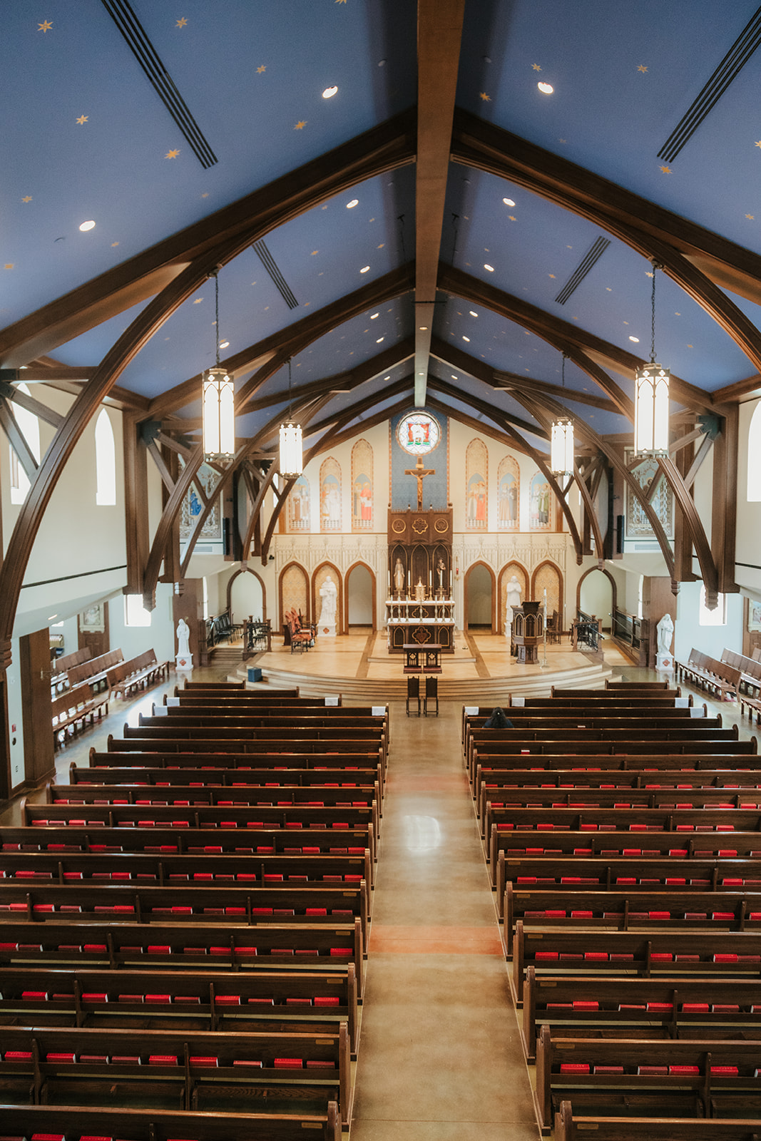 Interior view of a church with wooden pews, tall arched beams, hanging lights, and an altar at the front beneath a blue ceiling.