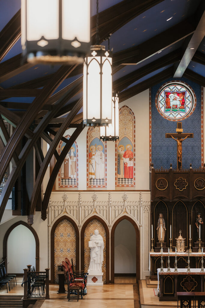 Interior of a church featuring arched wooden beams, stained glass window, religious statues, and an ornate altar with a large crucifix at St. Thomas More University Parish