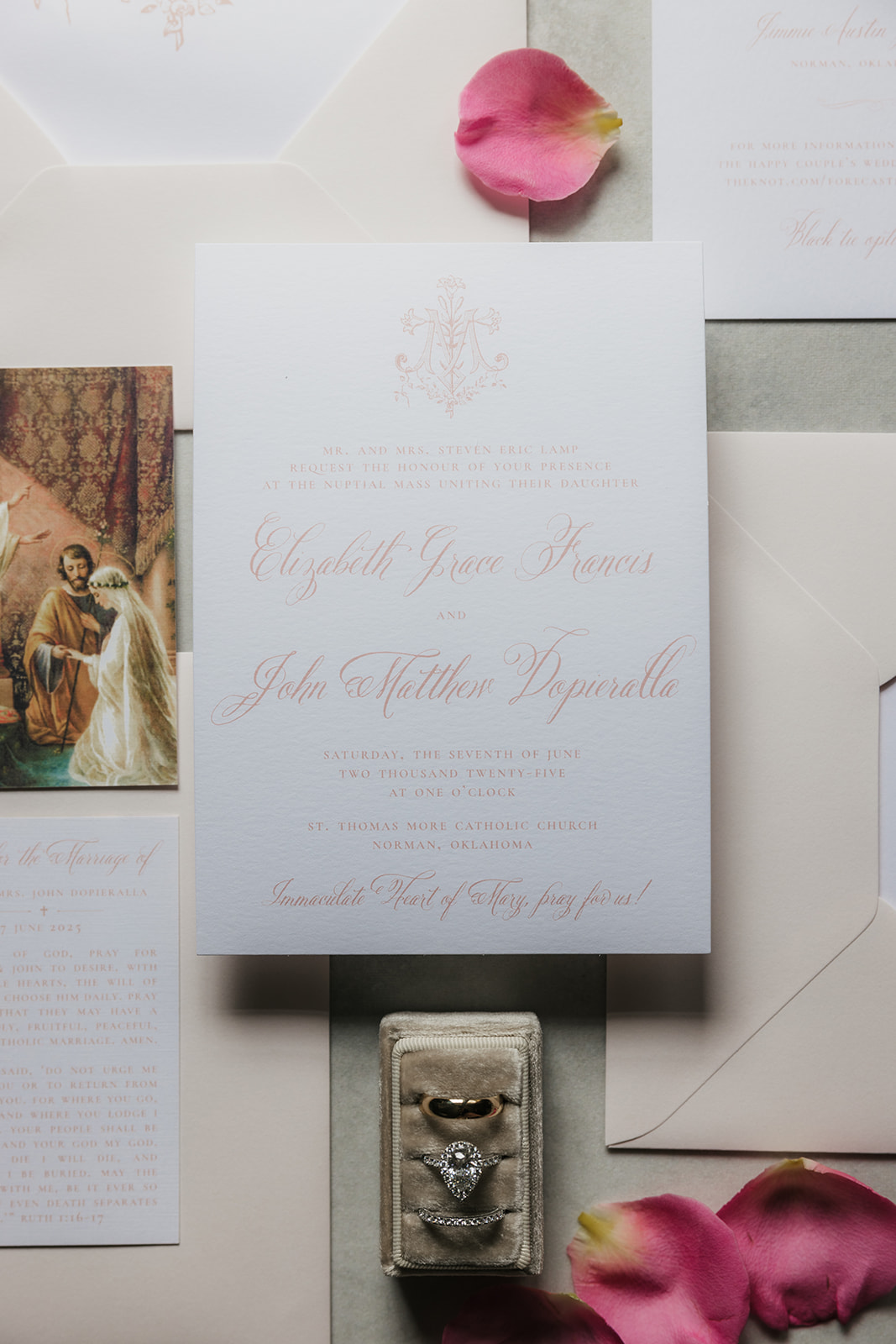 A wedding invitation card with pink script text lies on a blue surface, surrounded by pink rose petals and a beige ribbon.
