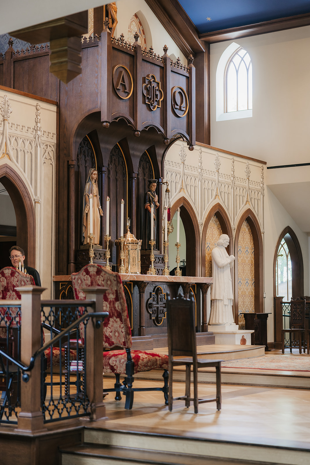 A church interior with a wooden altar, statue of a praying figure, candles, and religious decorations; arched windows and chairs are visible.