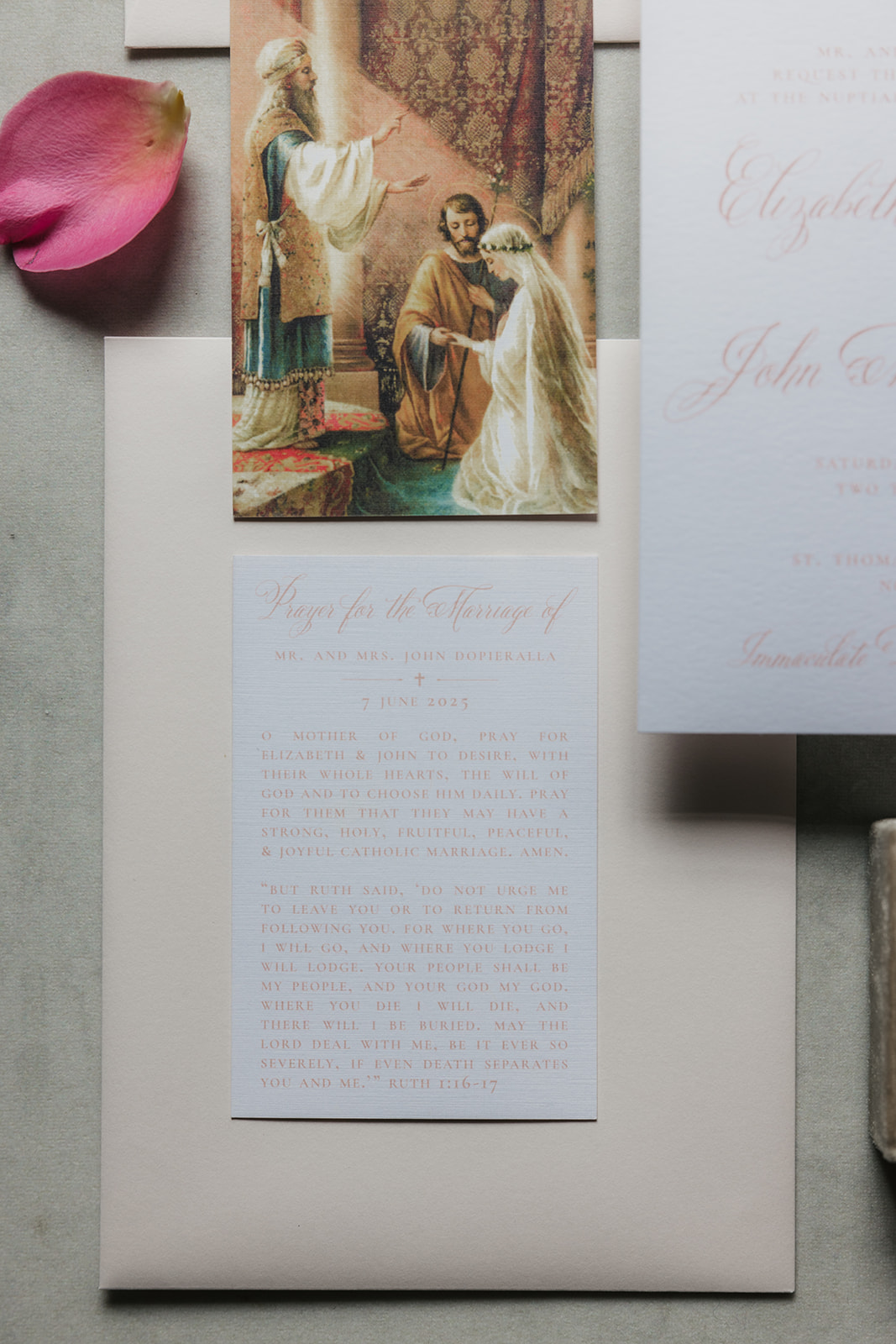 A wedding invitation card with pink script text lies on a blue surface, surrounded by pink rose petals and a beige ribbon.