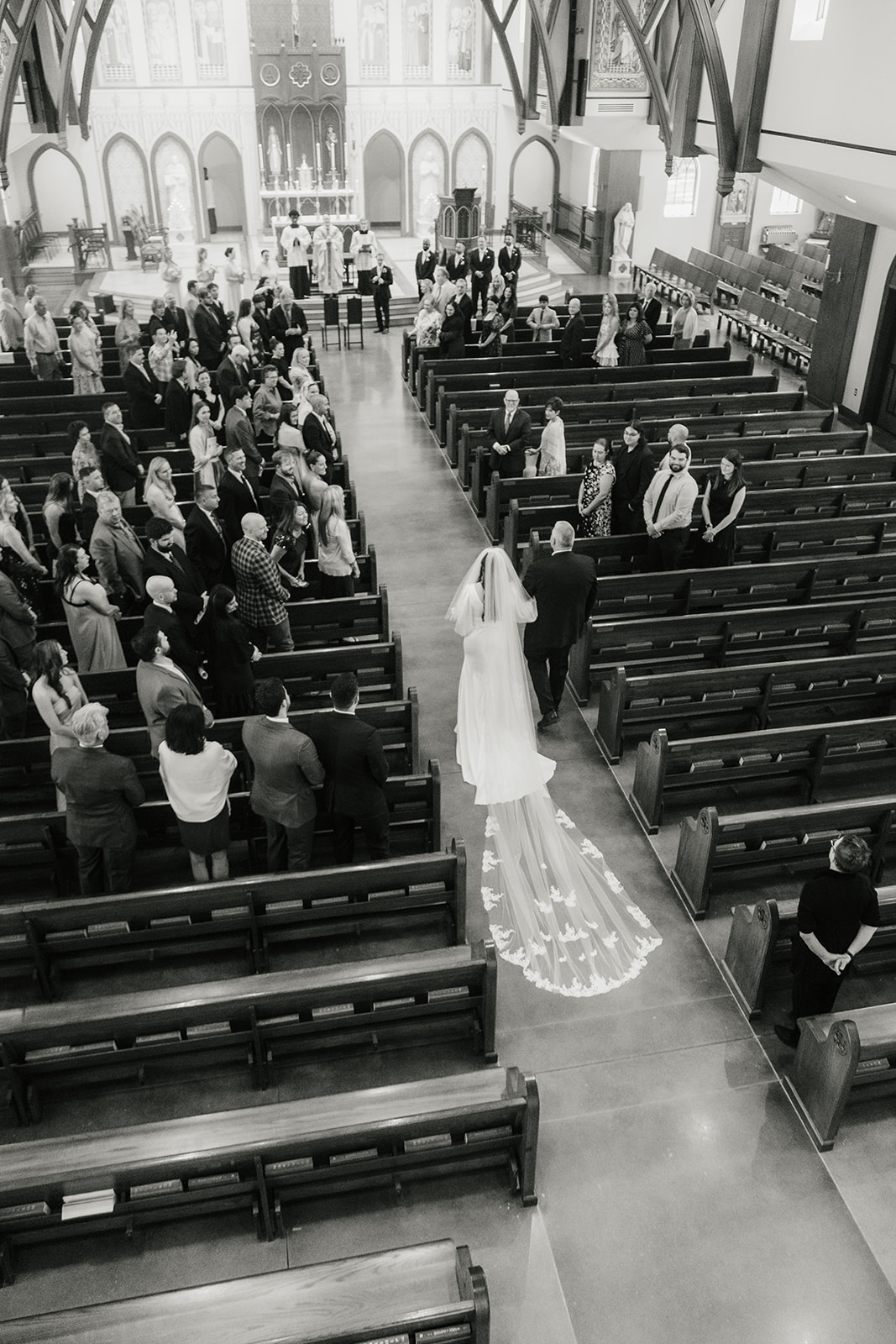A bride walks down the aisle of a church filled with seated and standing guests, viewed from above in black and white.