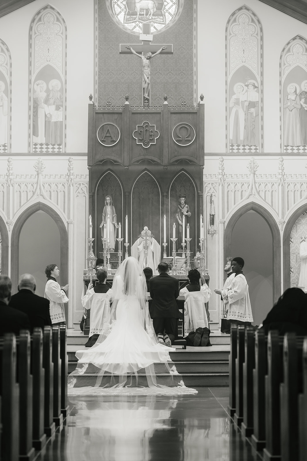 A bride and groom kneel at the altar during a wedding ceremony in a church, accompanied by clergy and altar servers for Wedding at St. Thomas More University Parish