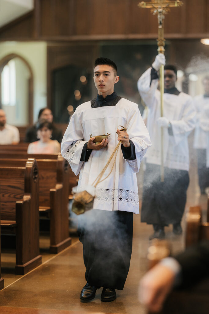 A young altar server in a white robe carries a censer, walking down the aisle of a church during a religious ceremony. Smoke from the censer is visible in the air for a Wedding at St. Thomas More University Parish