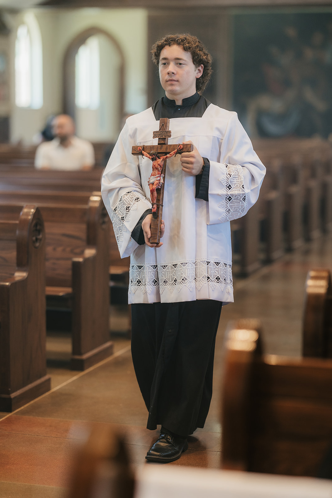 A person in religious vestments walks down a church aisle holding a crucifix, with wooden pews and stained glass windows in the background.