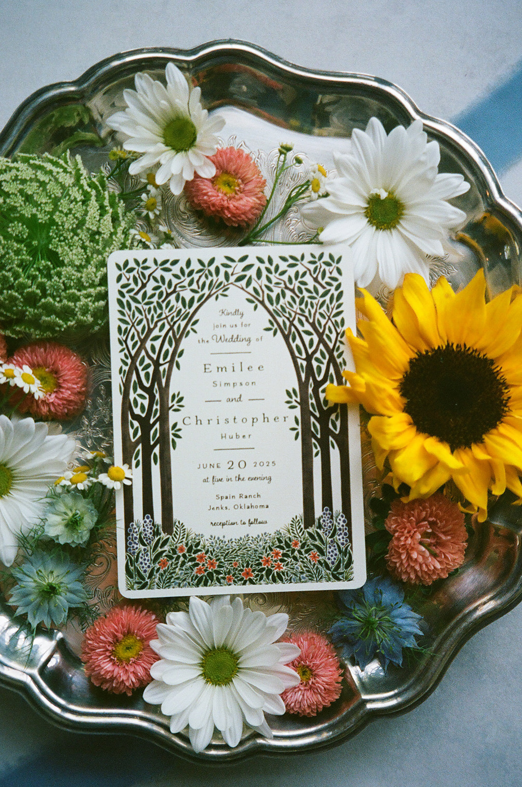 A decorative card with a tree design sits on a silver tray surrounded by assorted flowers, including a sunflower, daisies, and pink blooms.