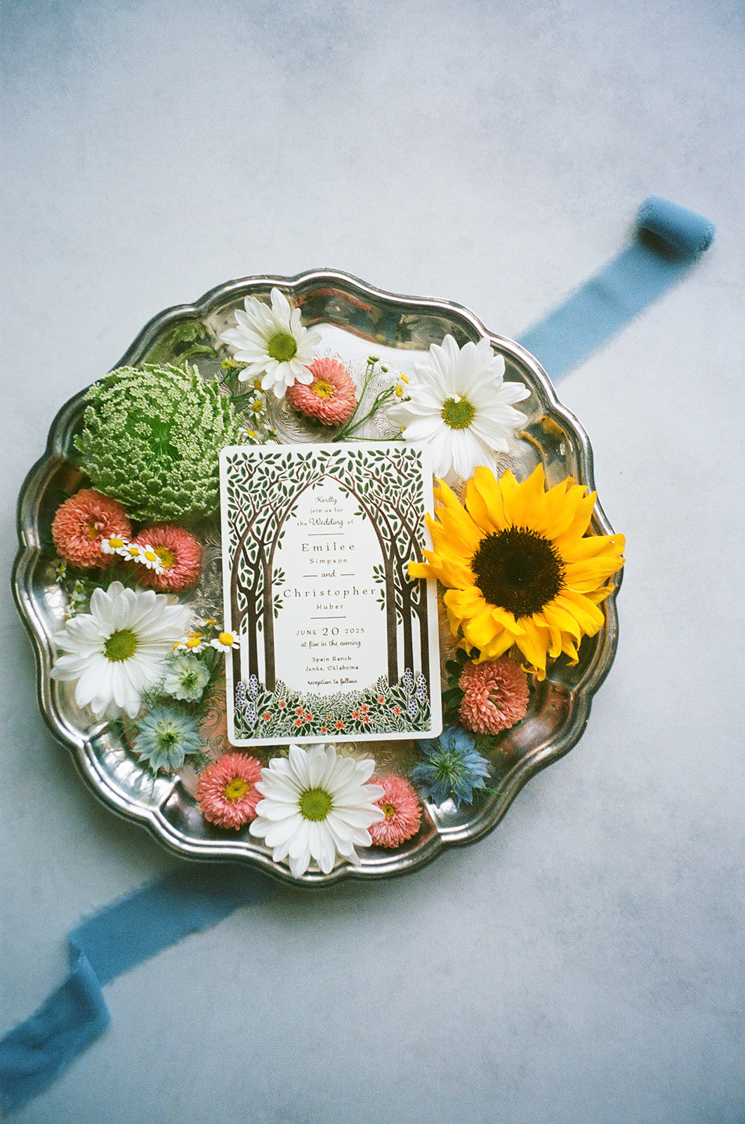 A decorative card with a tree design sits on a silver tray surrounded by assorted flowers, including a sunflower, daisies, and pink blooms.