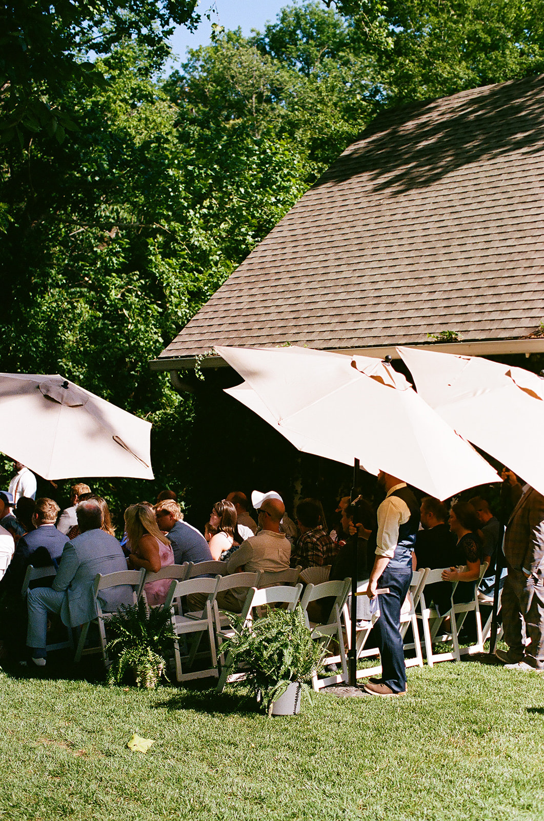 People sit on white chairs under large umbrellas at an outdoor event next to a building, surrounded by green trees and grass.