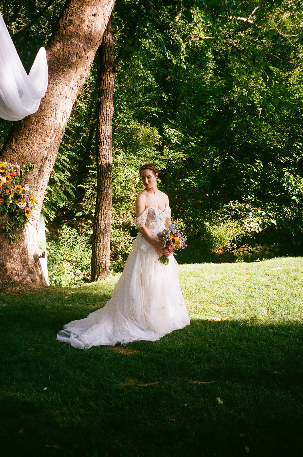 A bride and groom stand embracing in front of a garden with purple and pink flowers, with trees and a fence in the background.