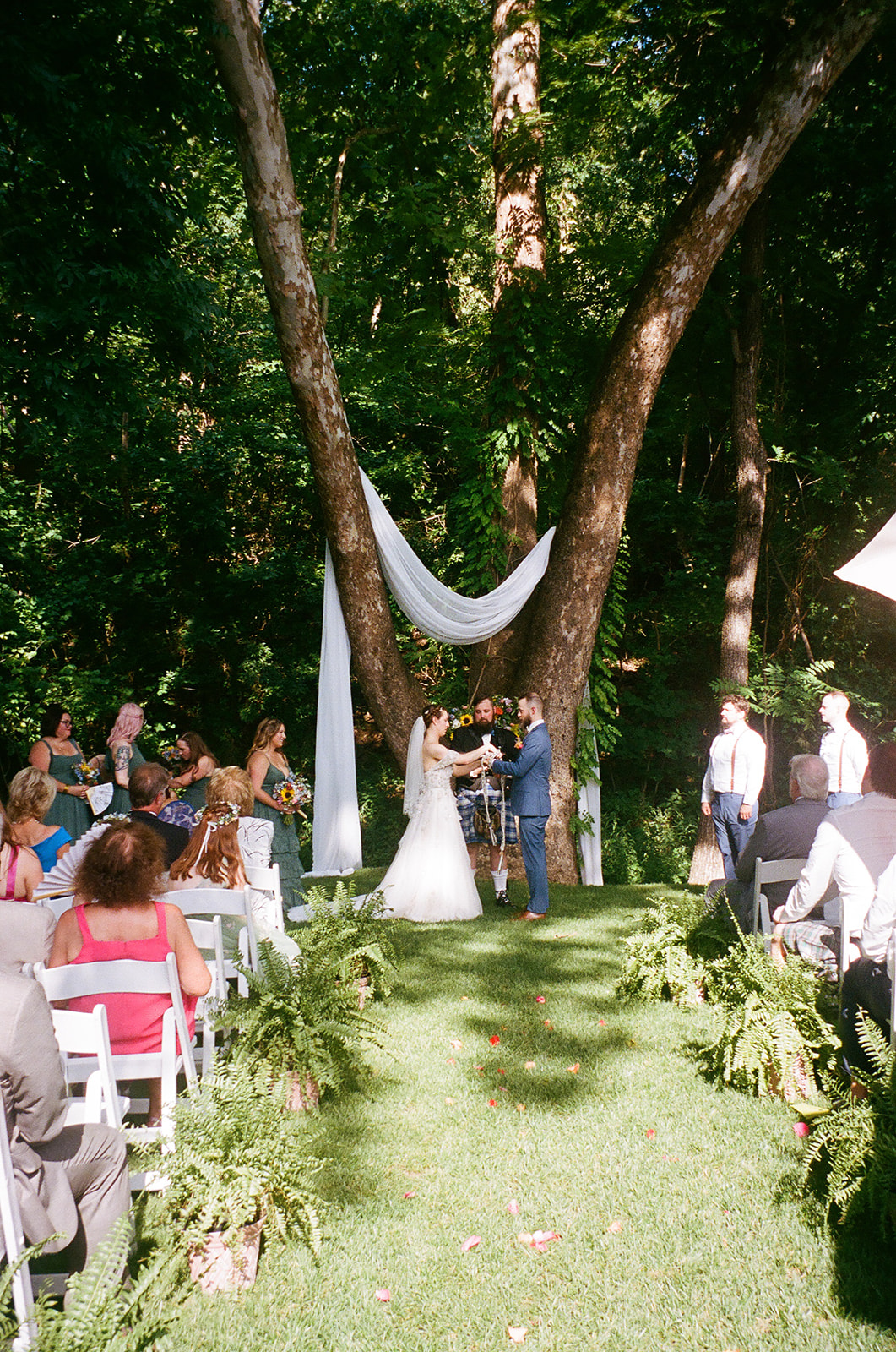 A bride and groom stand at an outdoor altar under trees, exchanging vows in front of seated guests on a sunny day for an outdoor wedding ceremony at  spain ranch