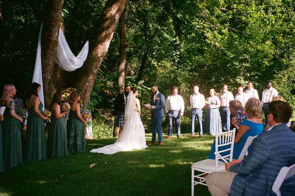 A bride and groom stand at an outdoor altar under trees, exchanging vows in front of seated guests on a sunny day for an outdoor wedding ceremony at  spain ranch