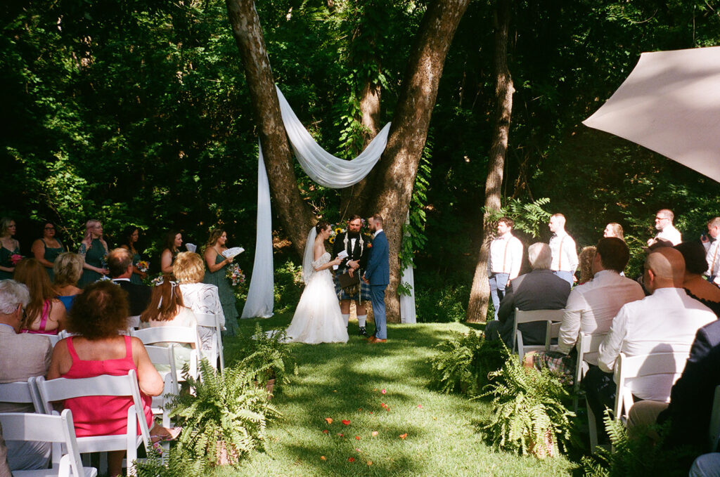 A bride and groom stand at an outdoor altar under trees, exchanging vows in front of seated guests on a sunny day for an outdoor wedding ceremony at  spain ranch