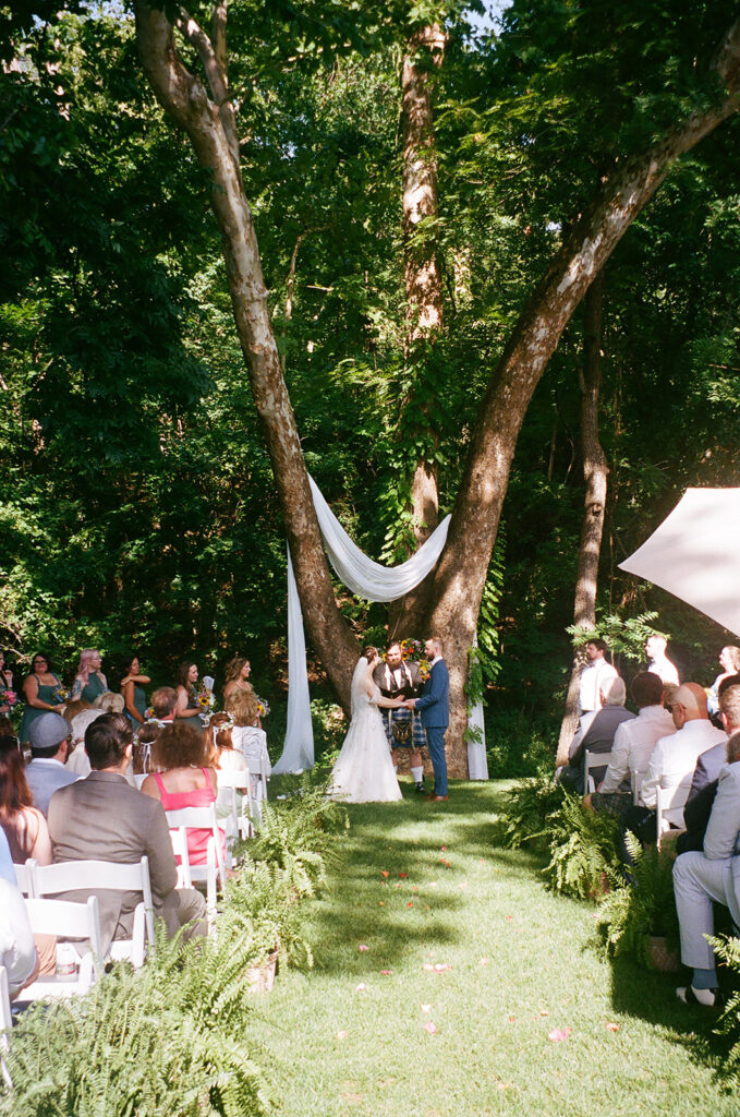 A bride and groom stand at an outdoor altar under trees, exchanging vows in front of seated guests on a sunny day for an outdoor wedding ceremony at  spain ranch
