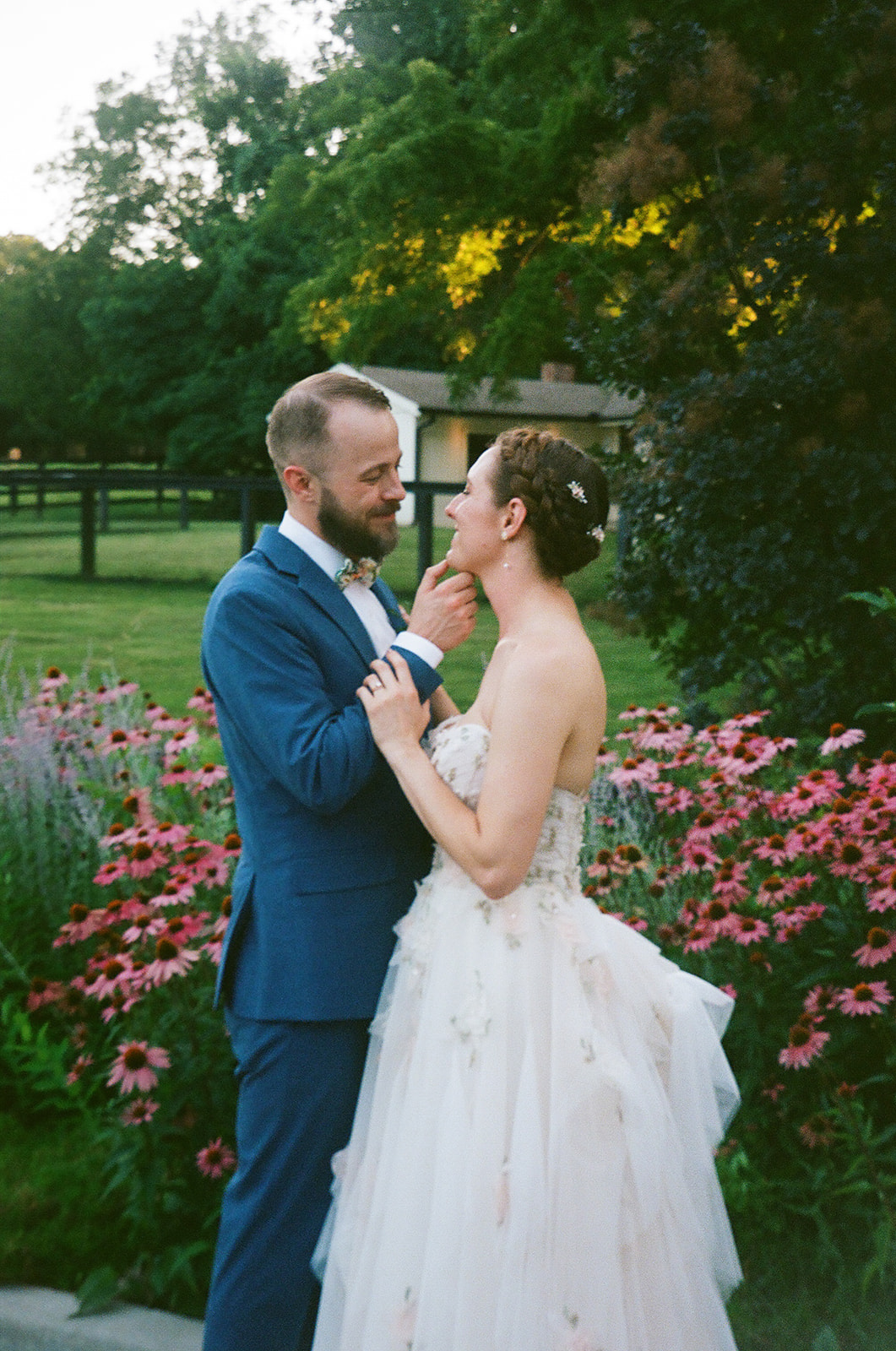 A couple dressed in wedding attire stands outdoors, surrounded by pink flowers and greenery, smiling at each other.