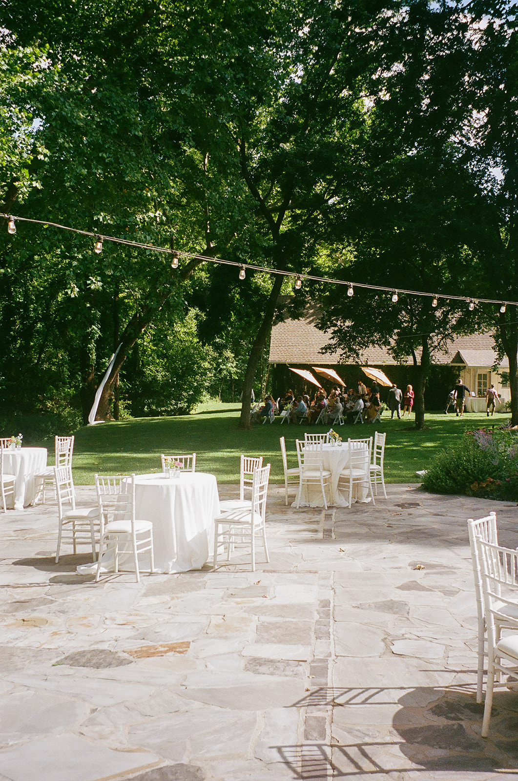 Outdoor patio with white tables and chairs on stone flooring, string lights overhead, and a wooden building with people in the background under green trees for a wedding at spain ranch