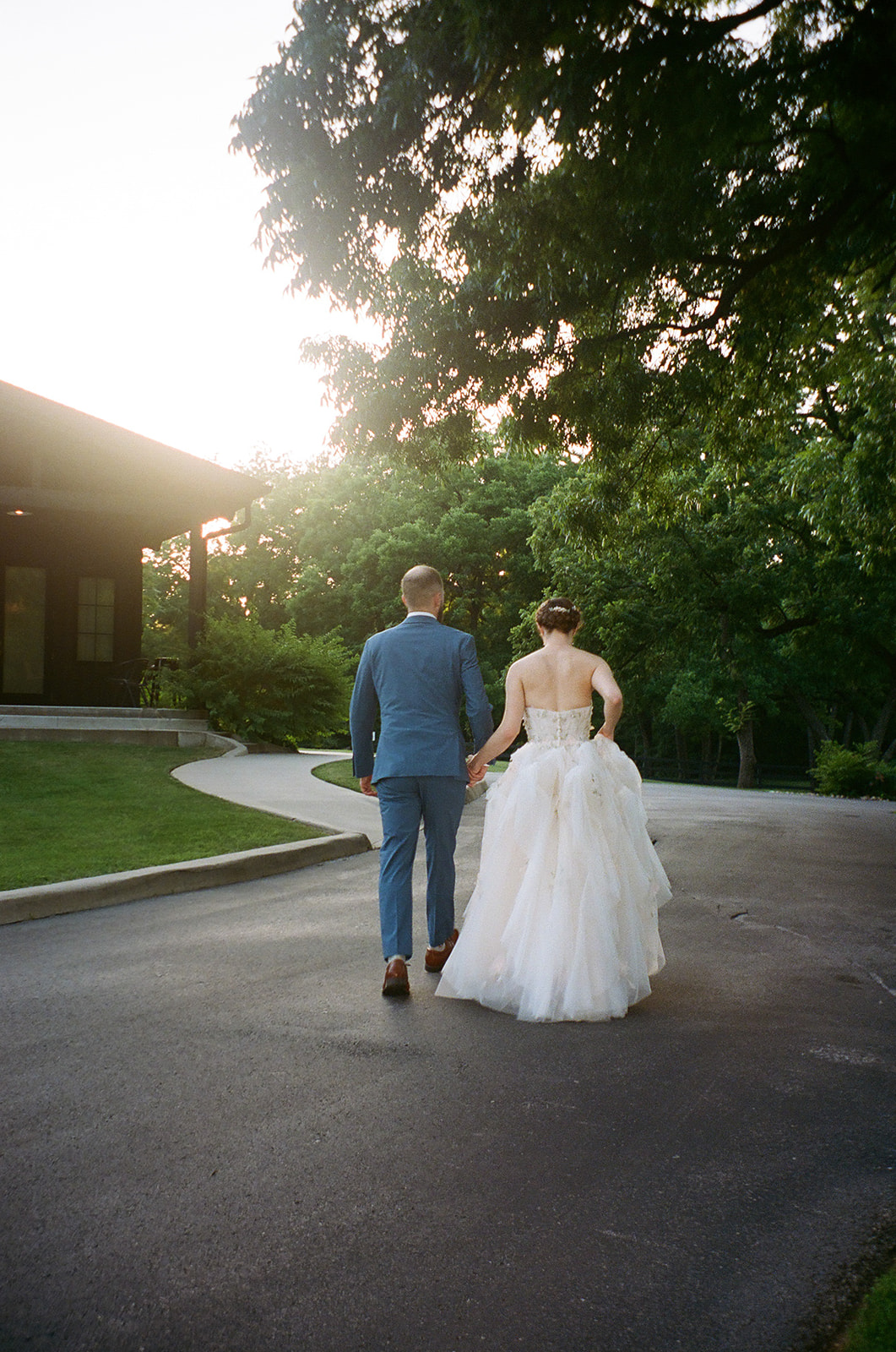 A couple dressed in wedding attire stands outdoors, surrounded by pink flowers and greenery, smiling at each other.