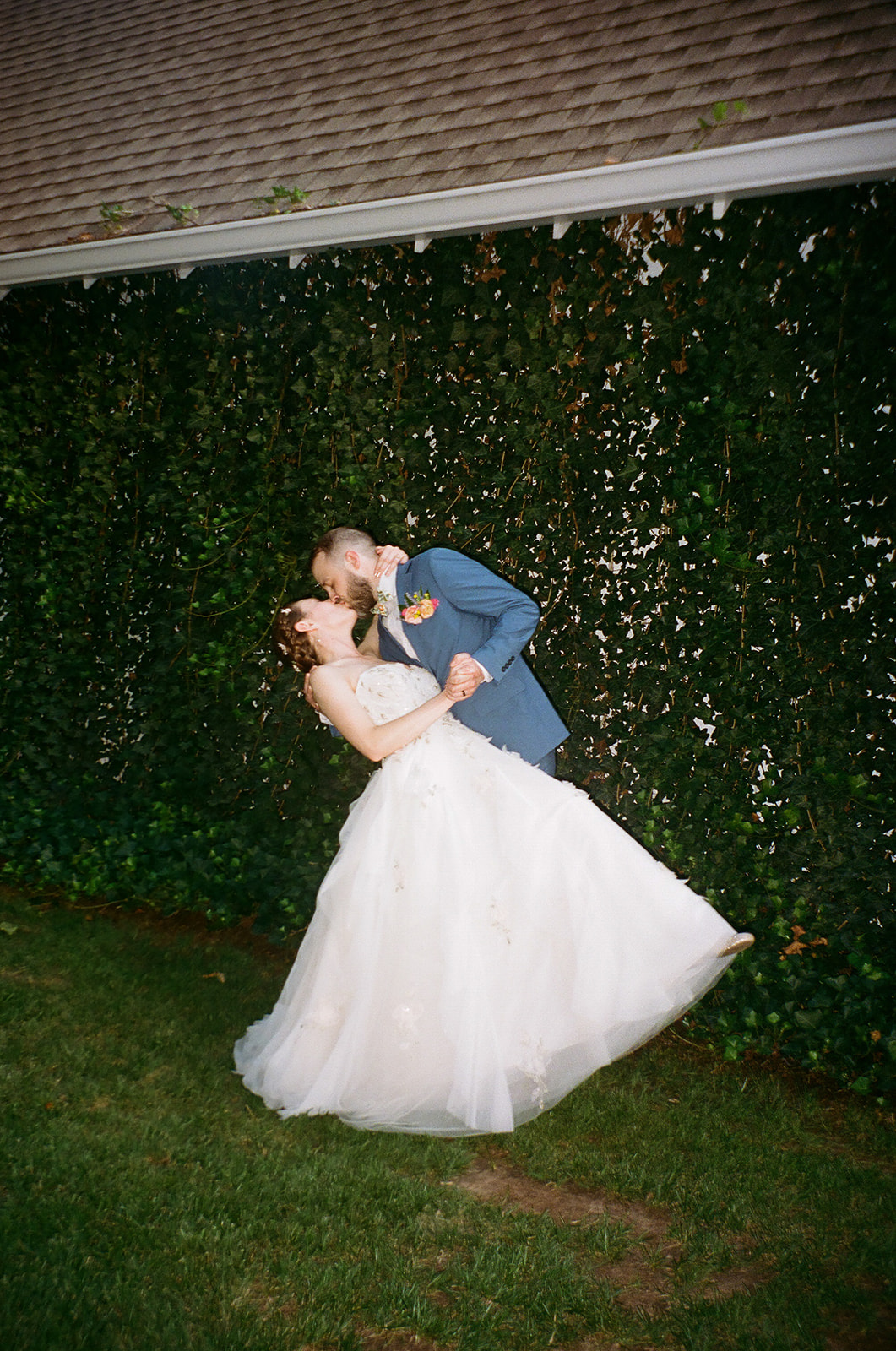 A couple dressed in wedding attire stands outdoors, surrounded by pink flowers and greenery, smiling at each other.