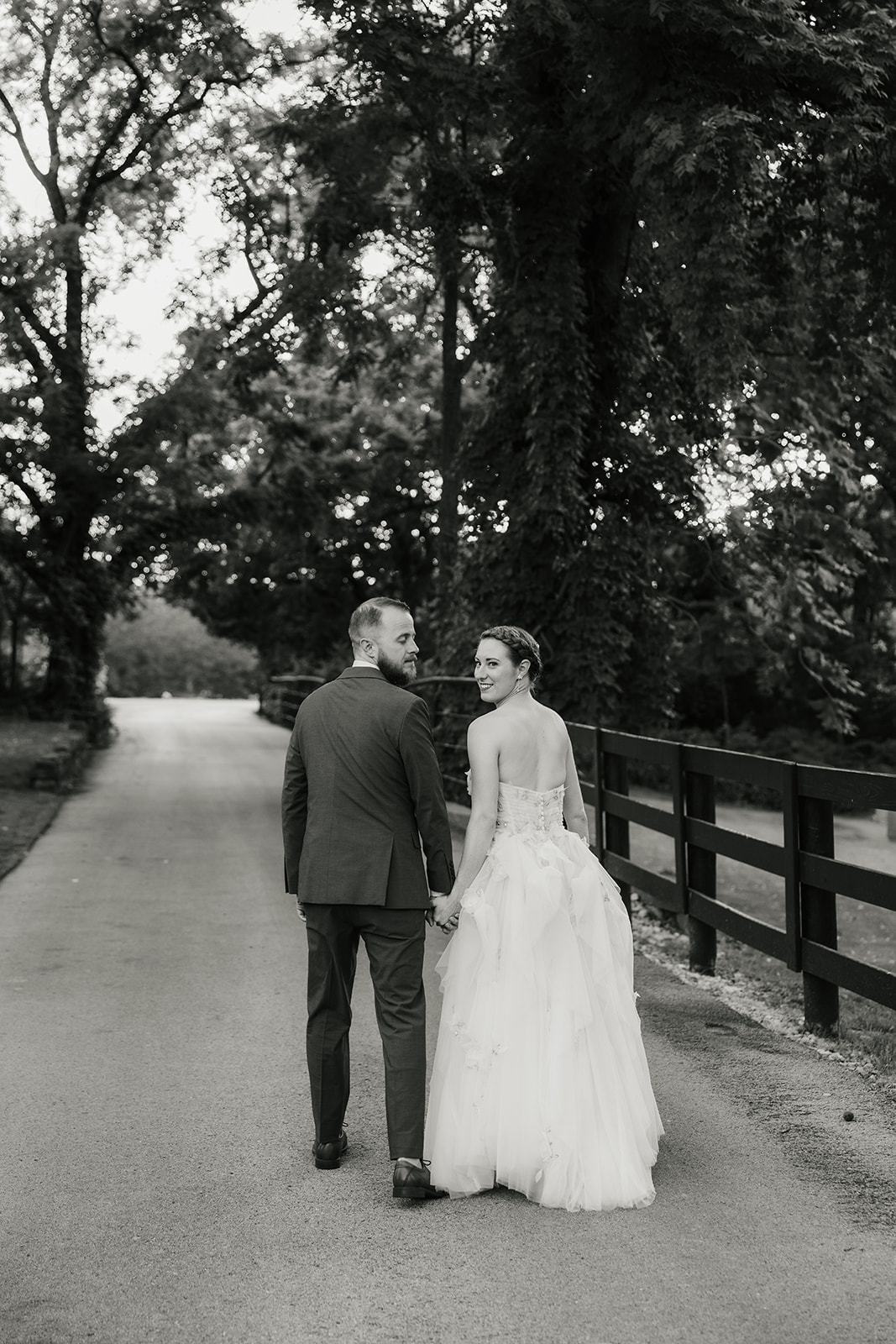 A bride in a white gown and a groom in a blue suit kiss while standing on pavement in front of a garden with pink flowers and green foliage.