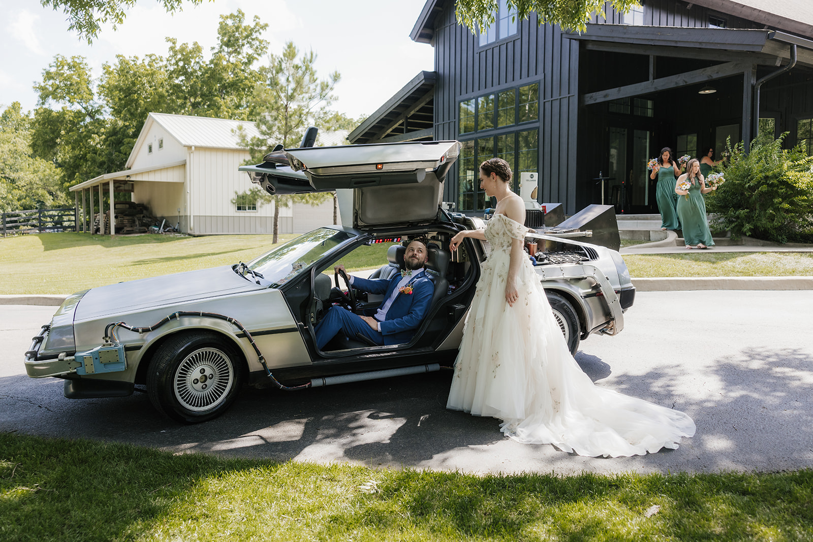 A bride in a white gown and a groom in a blue suit kiss while standing on pavement in front of a garden with pink flowers and green foliage.