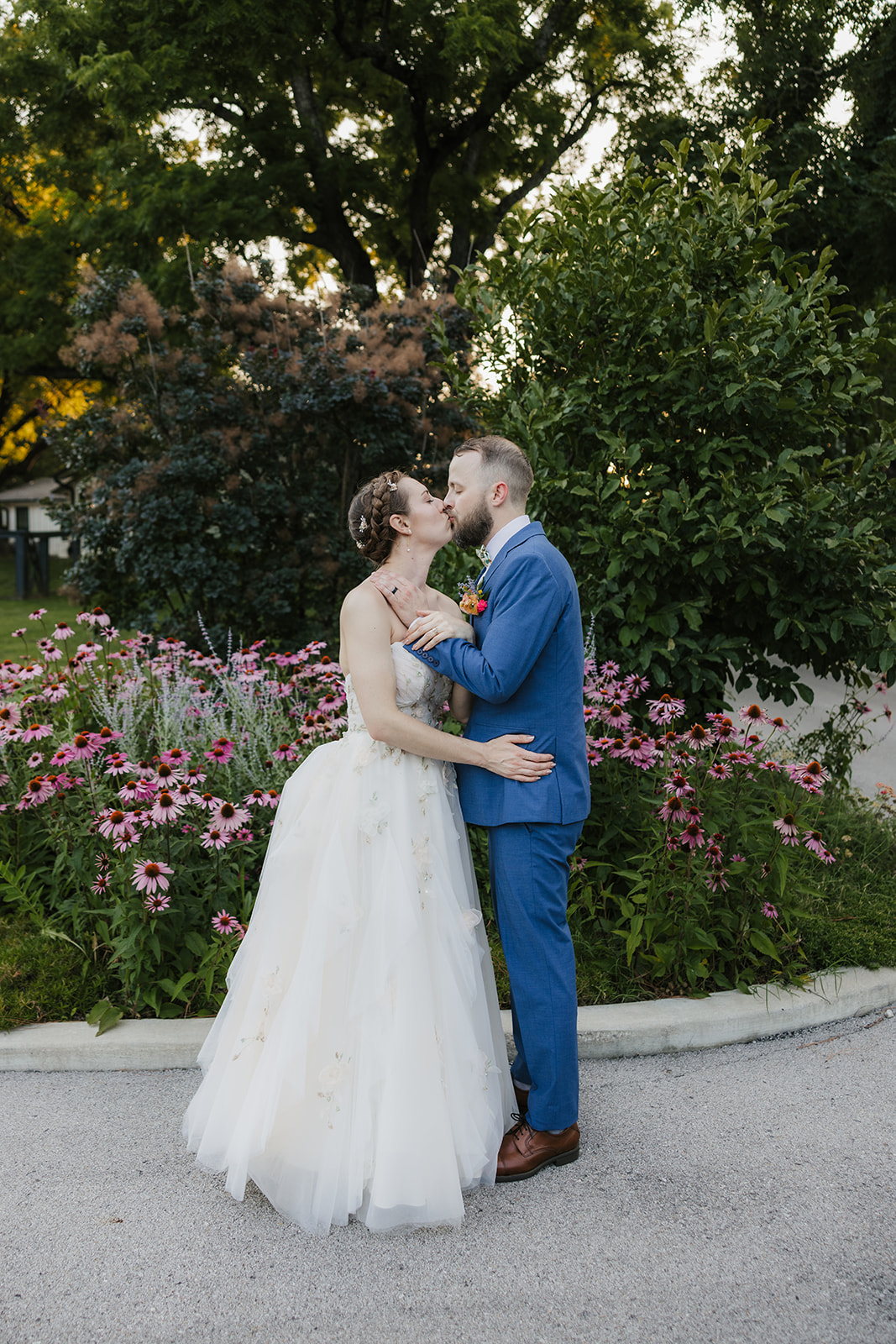 A bride in a white gown and a groom in a blue suit kiss while standing on pavement in front of a garden with pink flowers and green foliage.