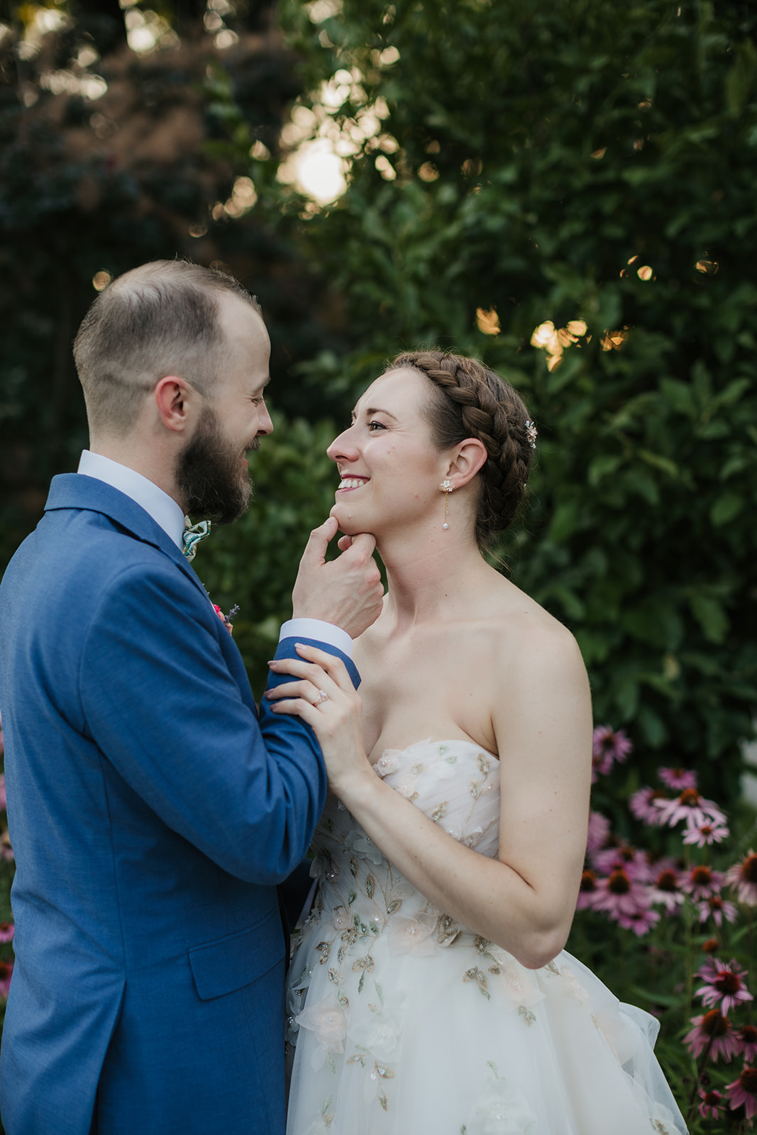 A bride in a white gown and a groom in a blue suit kiss while standing on pavement in front of a garden with pink flowers and green foliage.