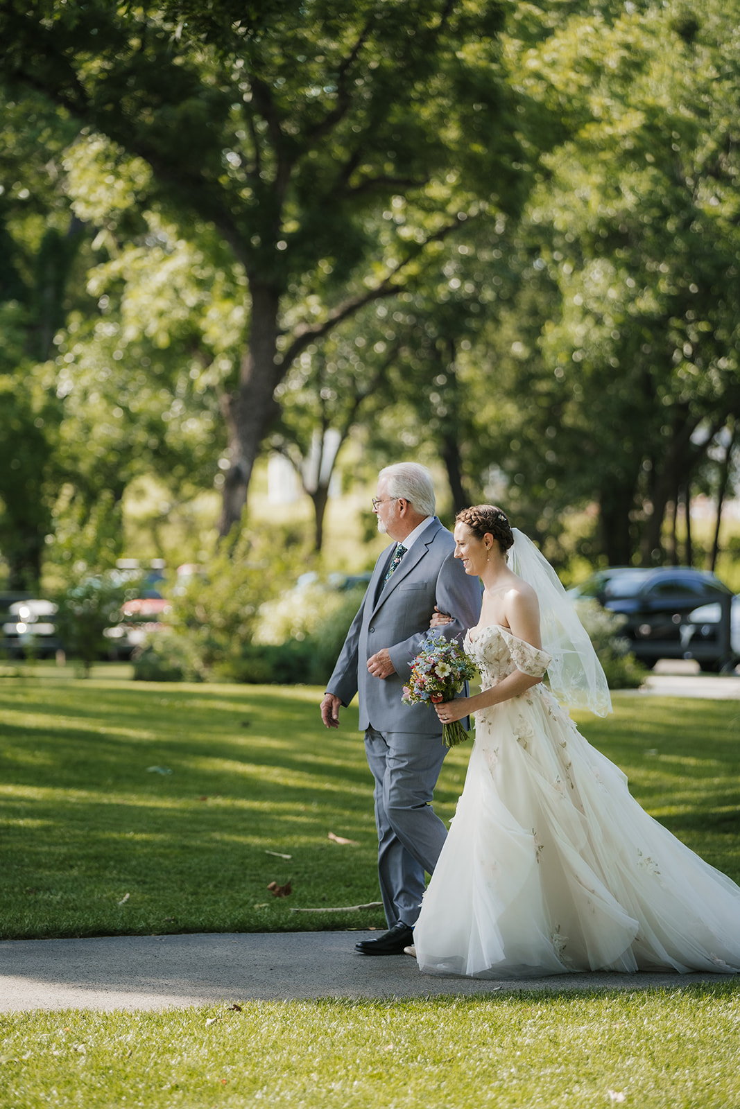 A bride in a white gown and veil walks outdoors on a path, accompanied by an older man in a gray suit, with trees and grass in the background.