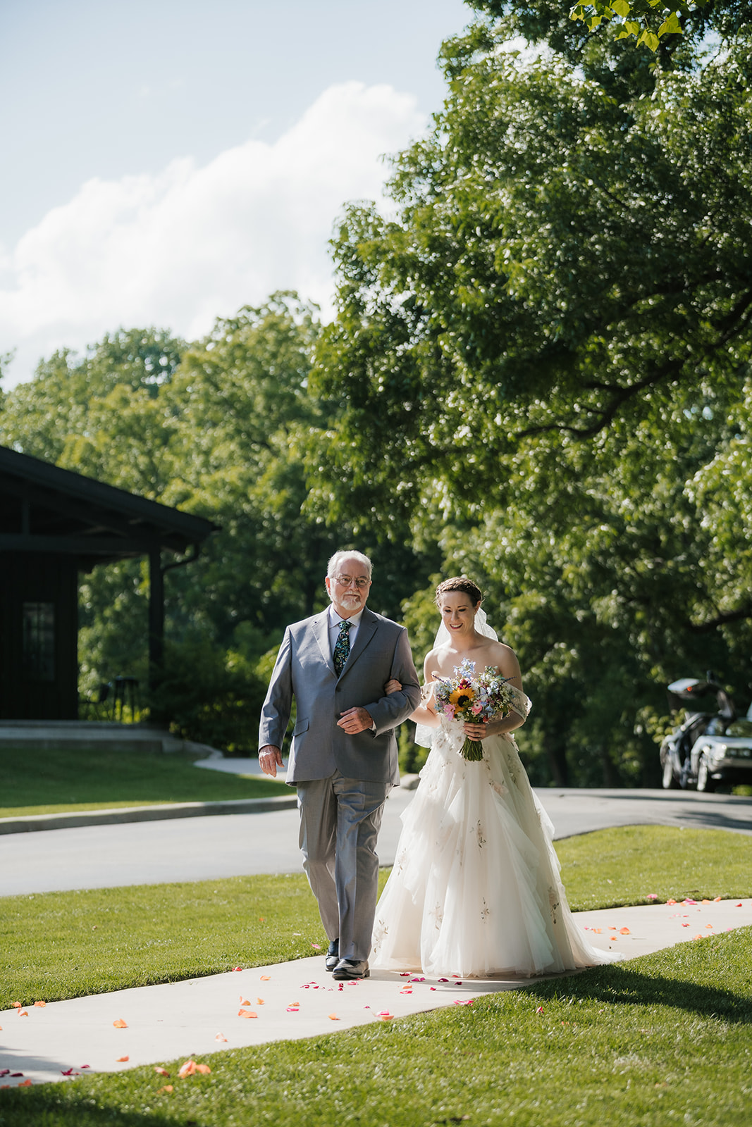A bride in a white gown and veil walks outdoors on a path, accompanied by an older man in a gray suit, with trees and grass in the background.