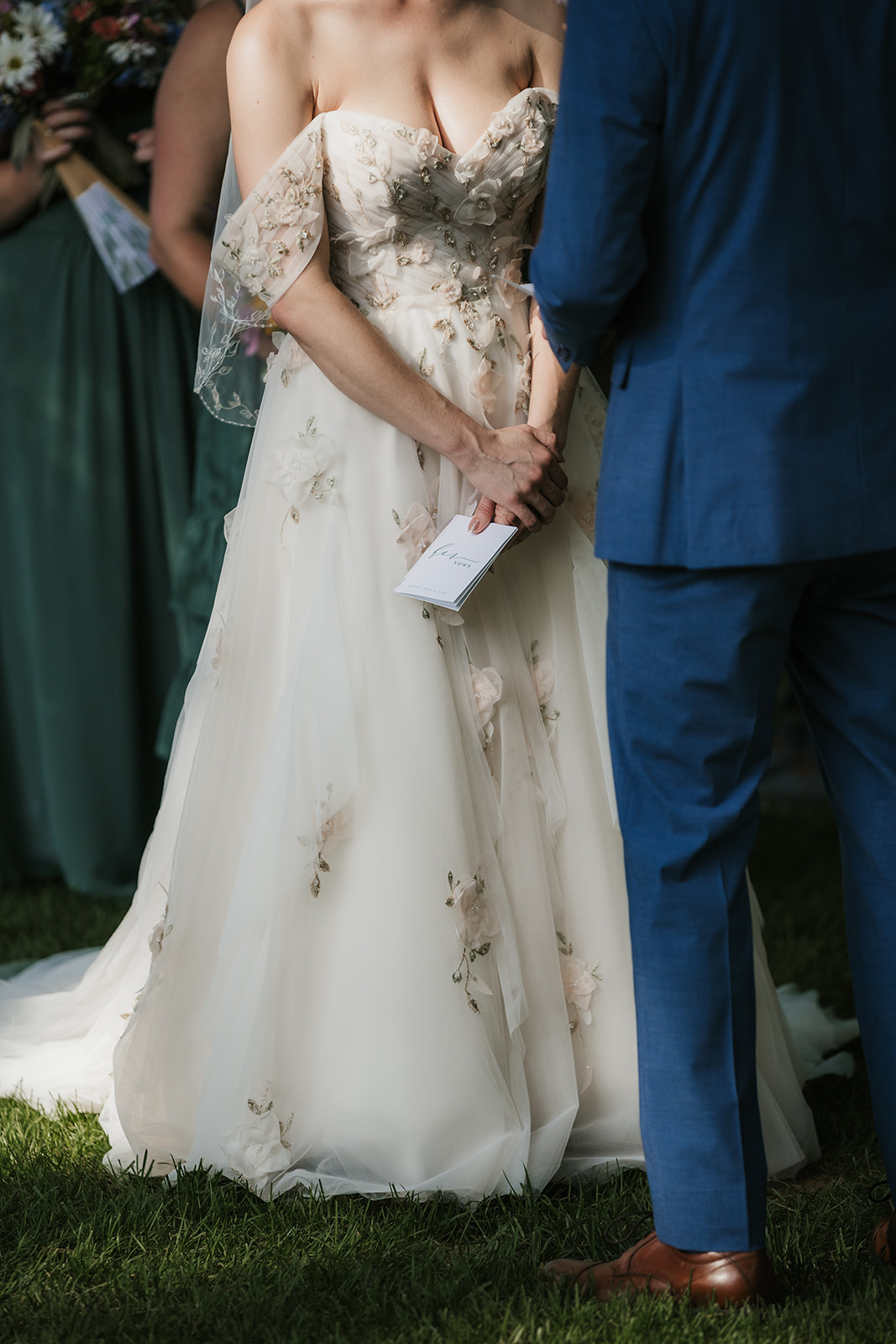 A bride in a white strapless gown and a groom in a blue suit stand facing each other outdoors, holding hands and reading vows.