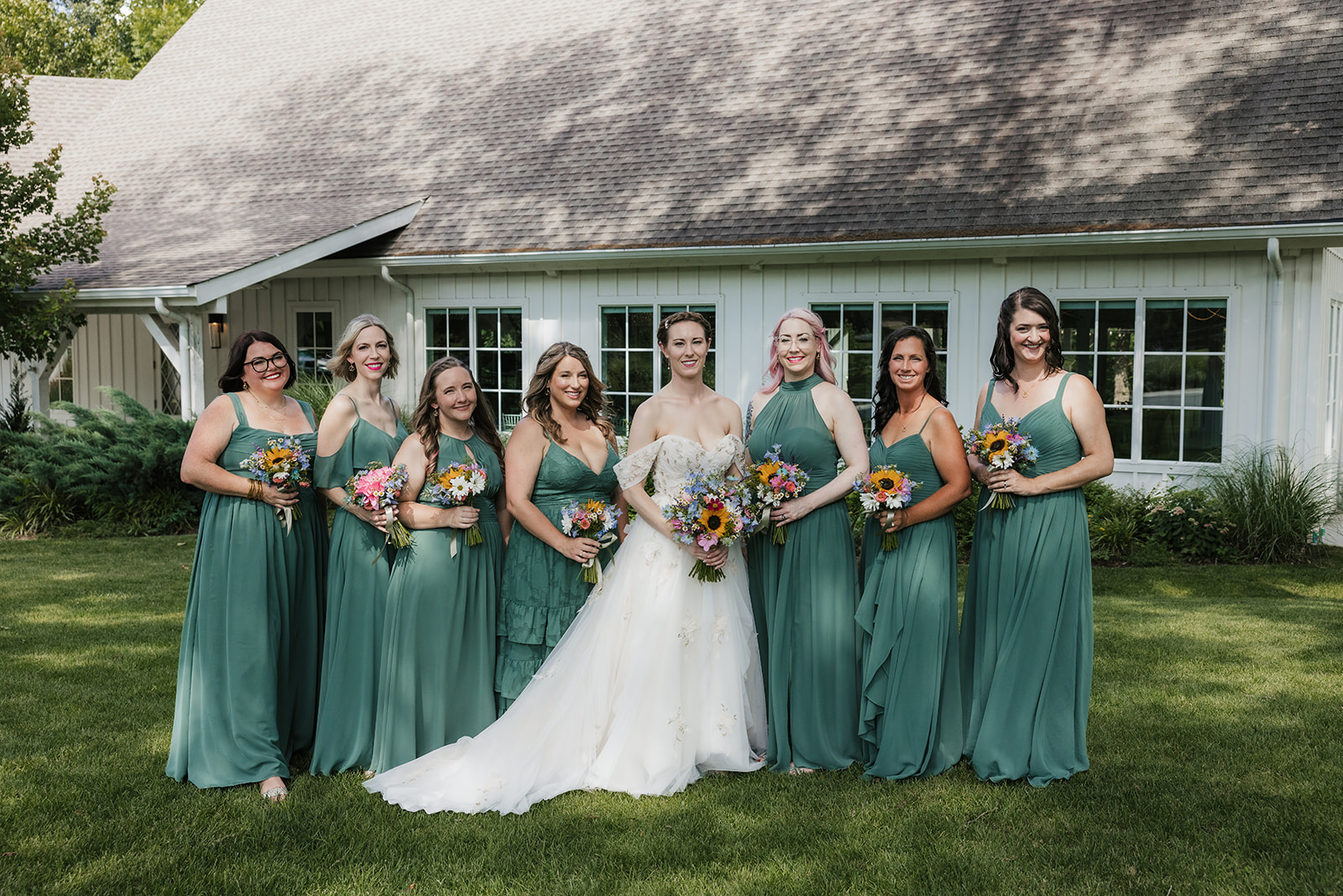 A bride in a white dress stands outdoors with seven bridesmaids in matching green dresses, all holding bouquets, in front of a white building for a wedding at spain ranch 