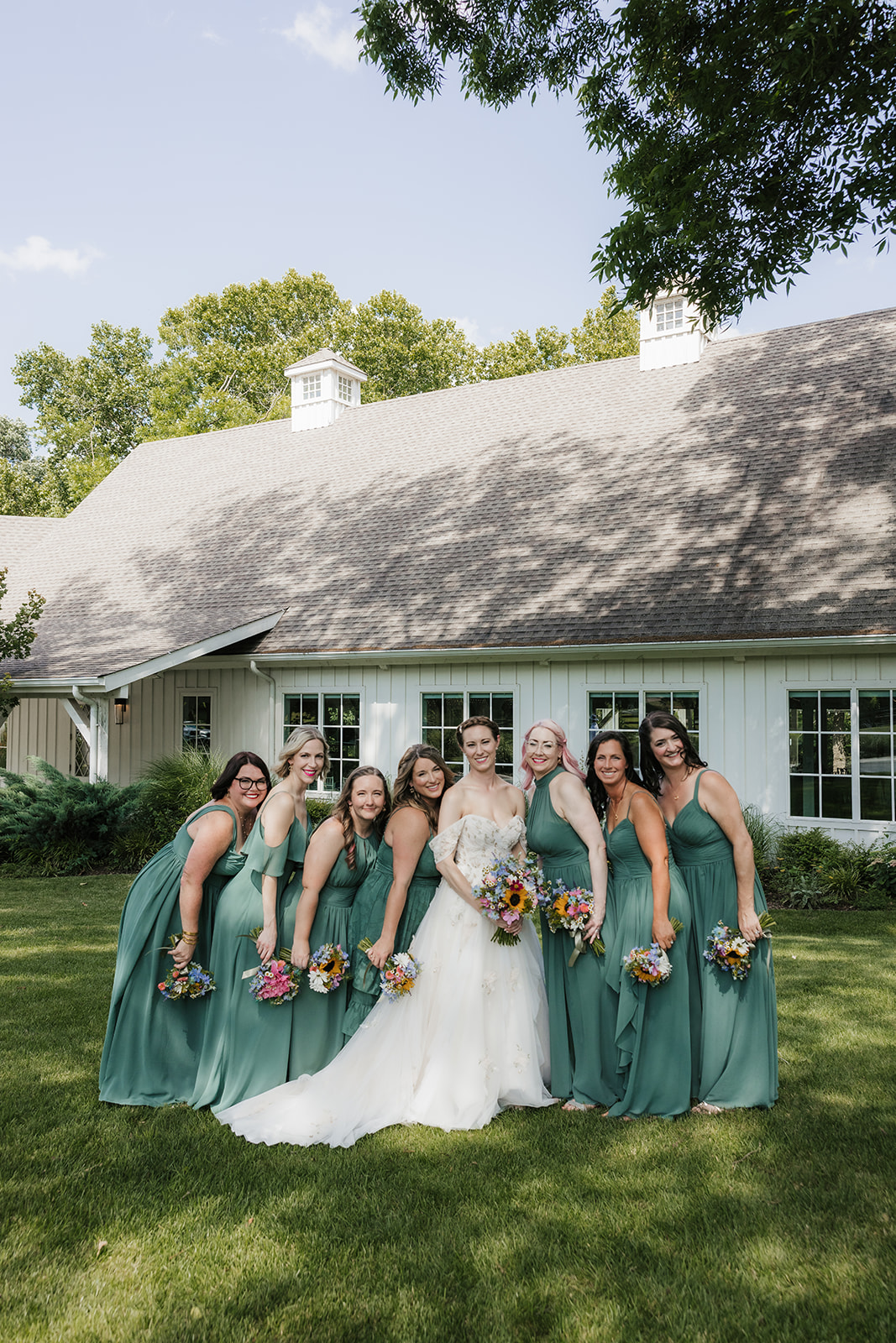 A bride in a white dress stands outdoors with seven bridesmaids in matching green dresses, all holding bouquets, in front of a white building for a wedding at spain ranch 