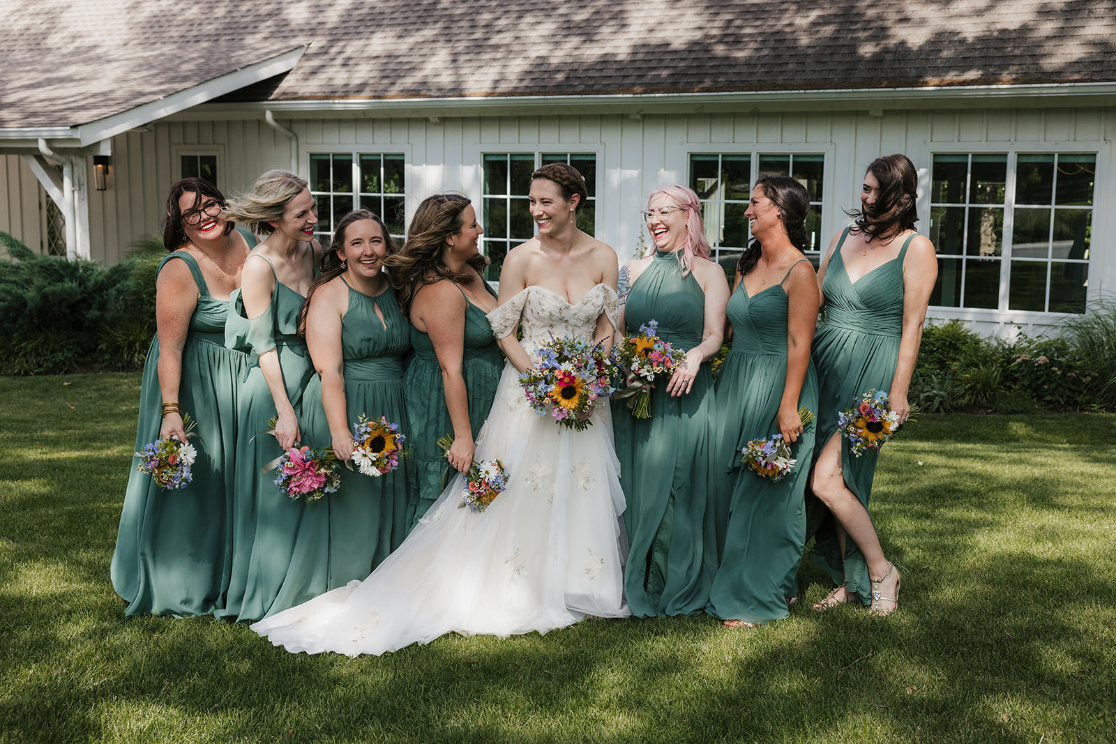 A bride in a white dress stands outdoors with seven bridesmaids in matching green dresses, all holding bouquets, in front of a white building for a wedding at spain ranch 