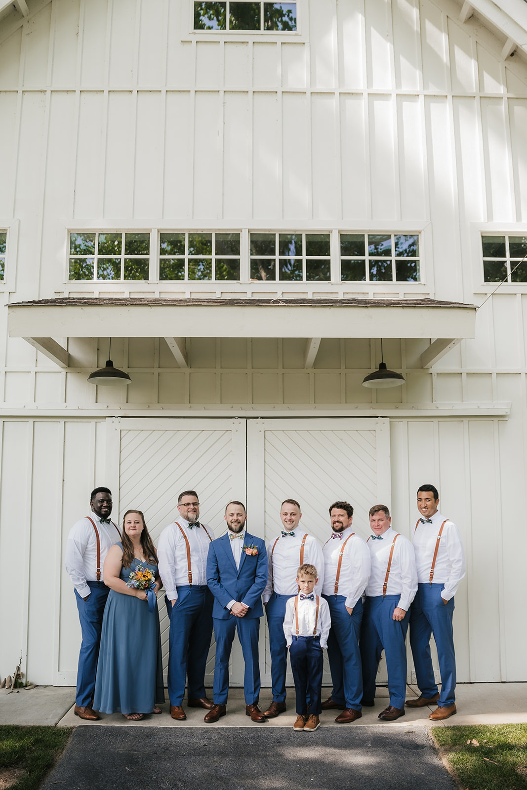 A wedding party poses in front of a white barn; the group includes men in blue pants and suspenders, a woman in a blue dress, and a child in matching attire.