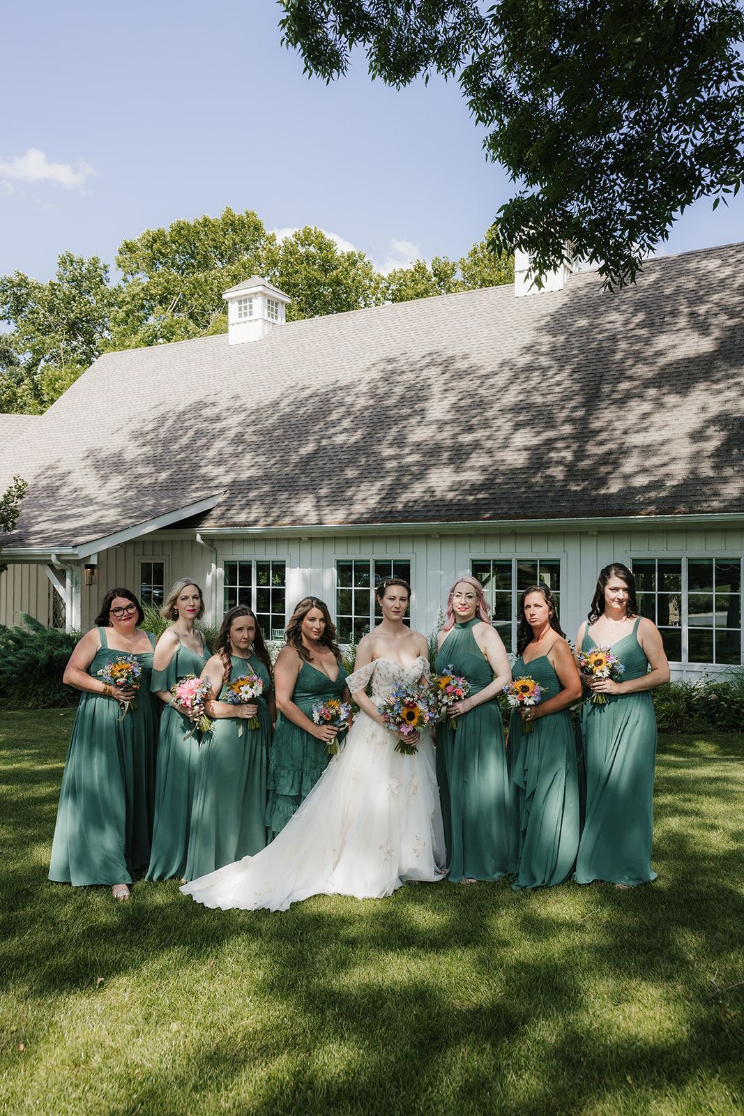 A bride in a white dress stands outdoors with seven bridesmaids in matching green dresses, all holding bouquets, in front of a white building for a wedding at spain ranch 