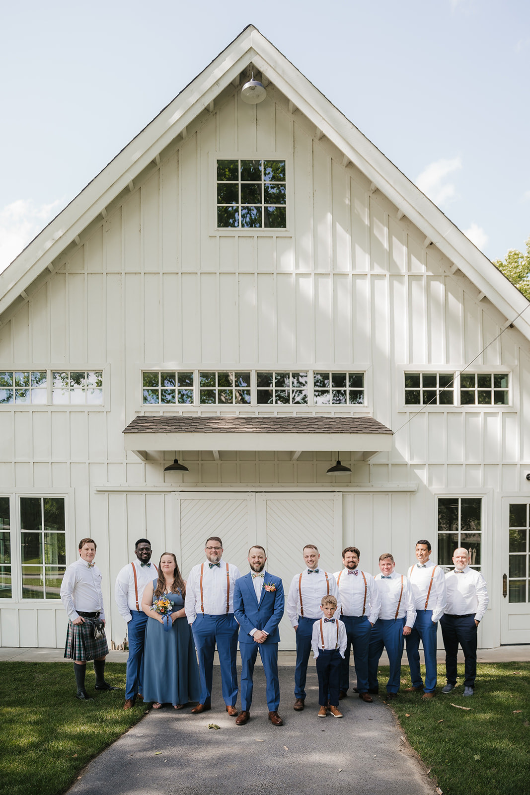 A wedding party poses in front of a white barn; the group includes men in blue pants and suspenders, a woman in a blue dress, and a child in matching attire.