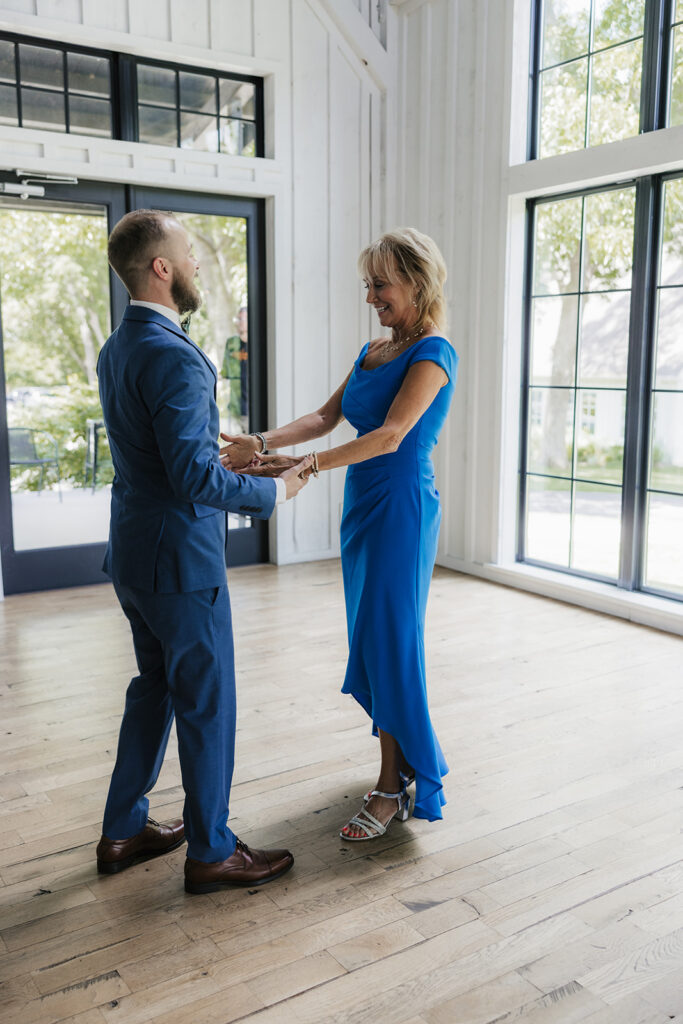 A man in a suit and a woman in a blue dress hold hands and smile at each other while standing in a bright room with large windows.